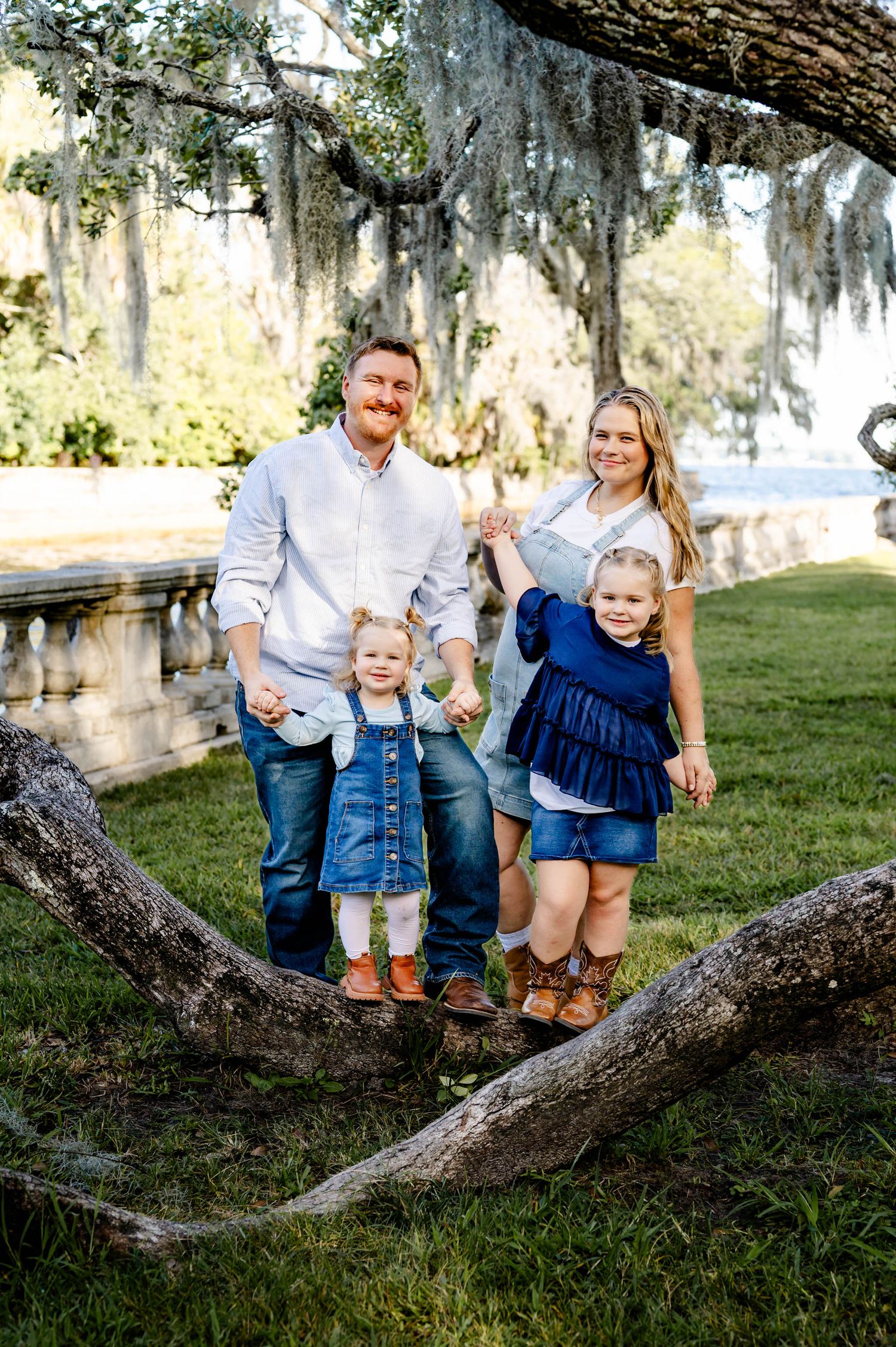 Family enjoying a sunny day together sitting on a large tree branch draped with Spanish moss in a park setting.