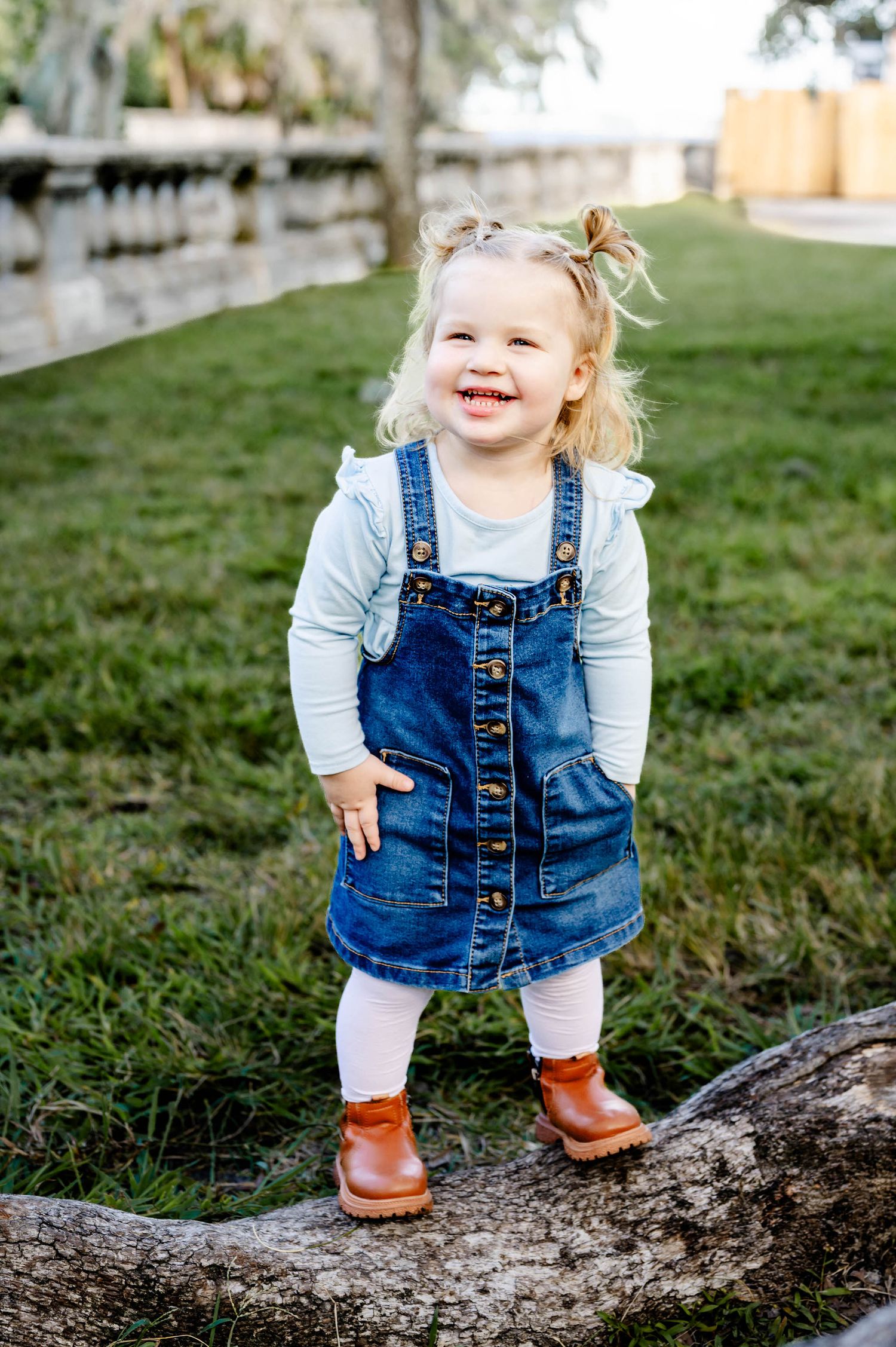 Small child in denim overalls and brown boots stands playfully on a fallen tree trunk in a grassy outdoor setting.