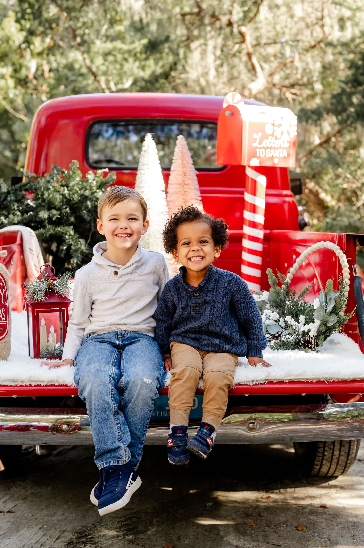Two children sit together in the back of a vintage red pickup truck decorated with Christmas trees and snow.
