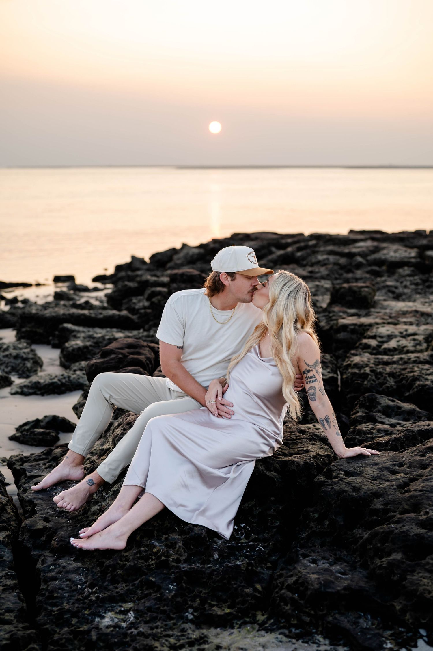 Couple sharing intimate moment while sitting on rocky coastline during sunrise maternity session.