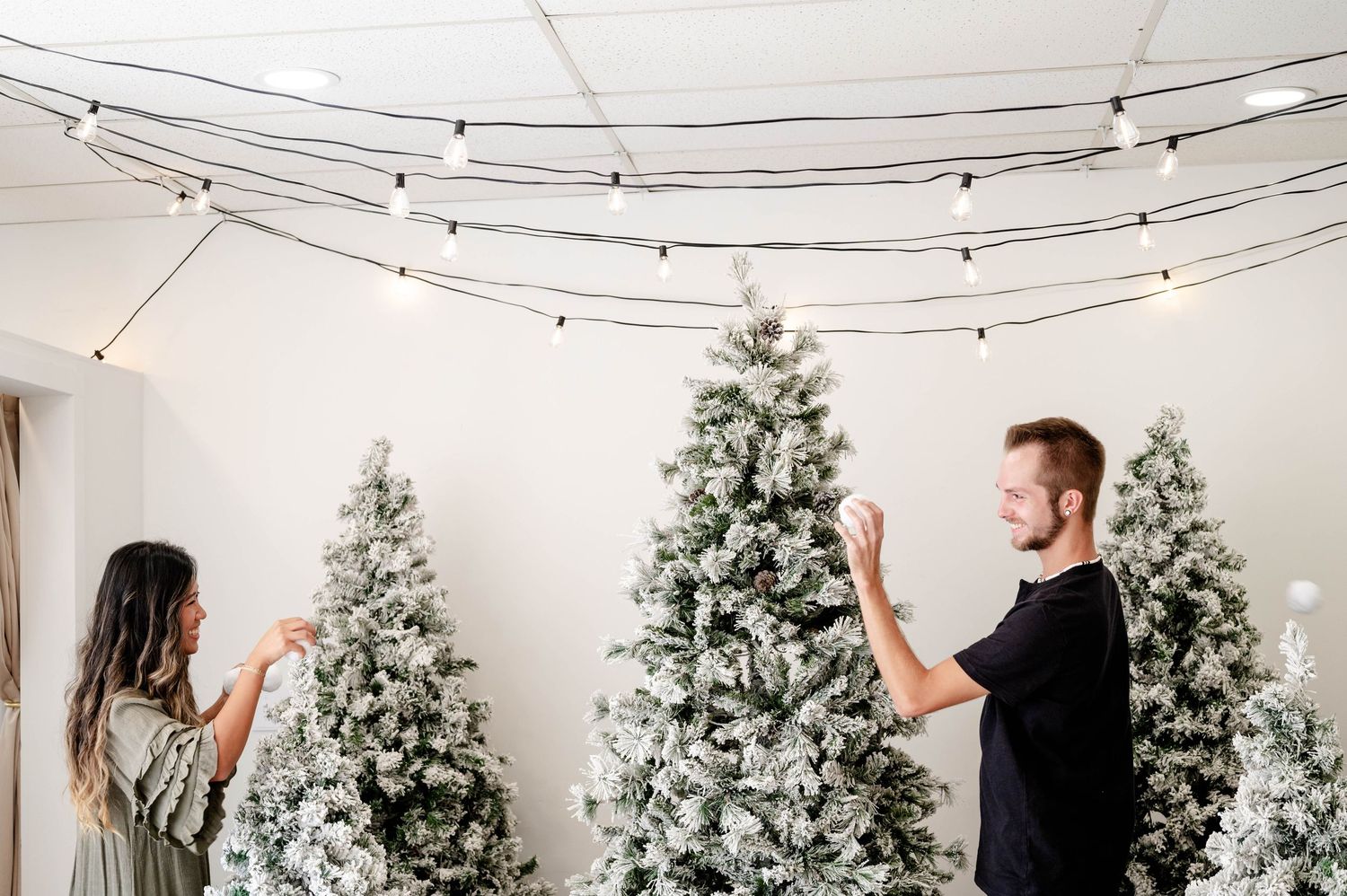 Two people decorating with string lights around flocked Christmas trees in a white room.