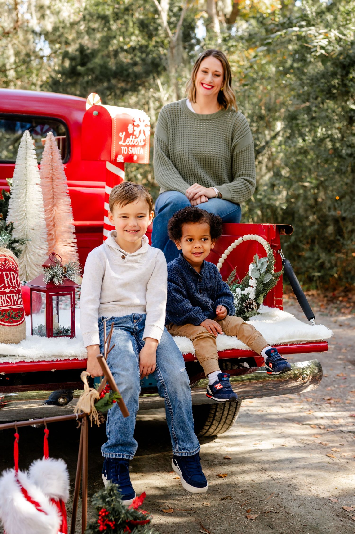 Family poses for Christmas photos on vintage red truck with white bottle brush trees and holiday decorations.