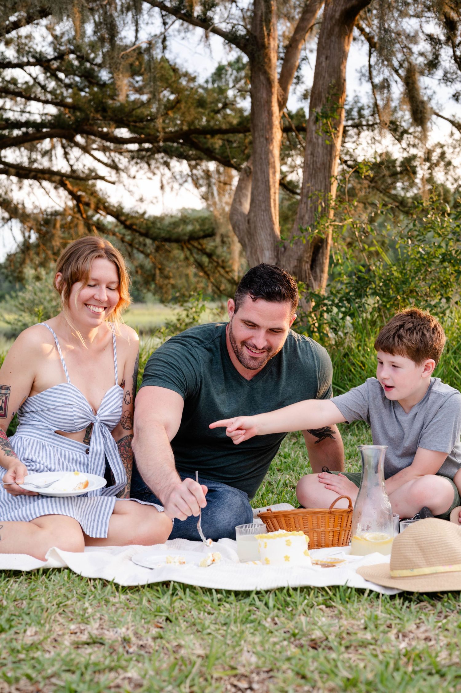 A family enjoying a picnic outdoors on a blanket with snacks and refreshments under tall trees.