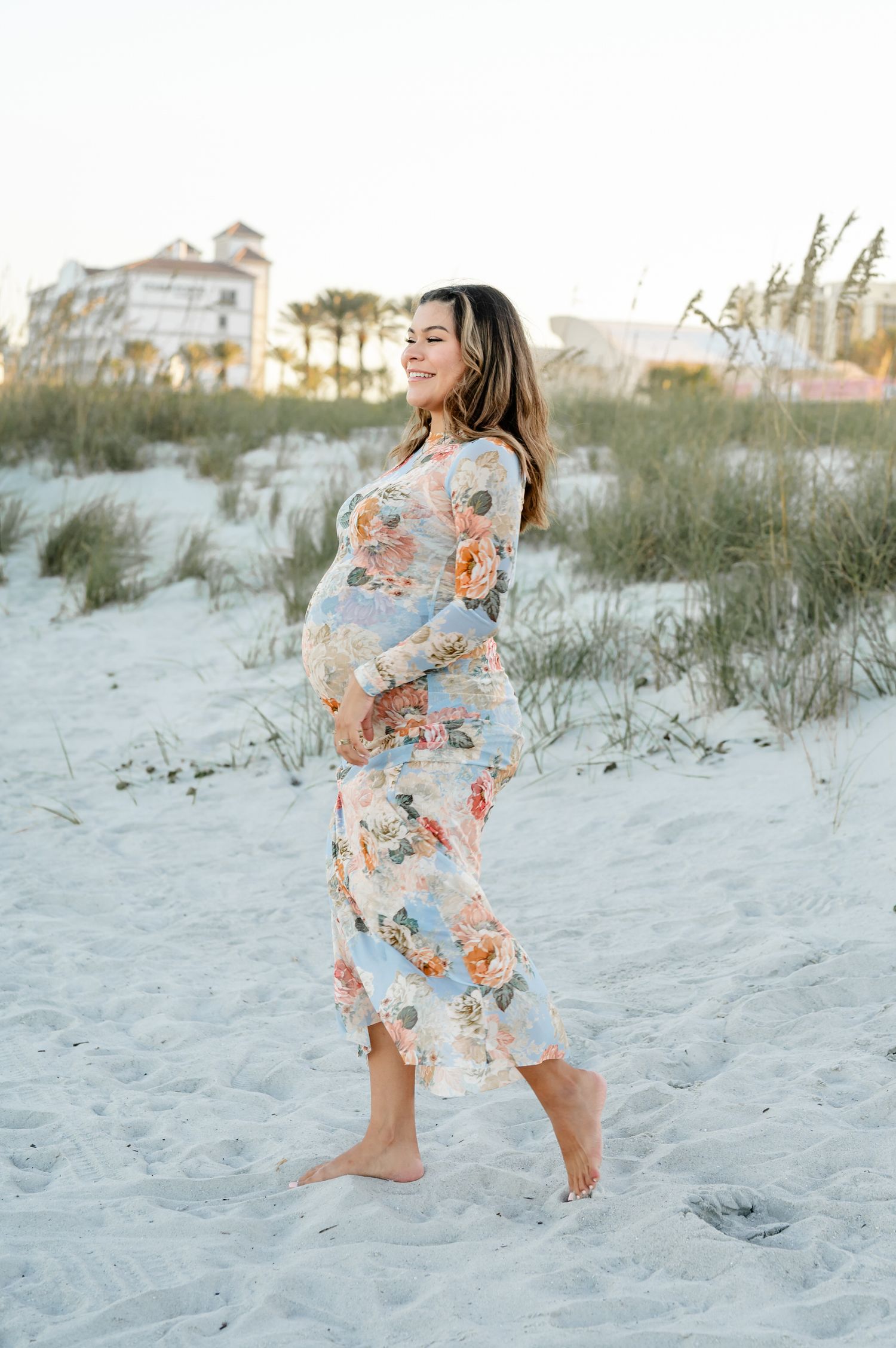 Pregnant woman in floral maxi dress smiling on white sand beach at sunset during a maternity photoshoot in Jacksonville, FL.