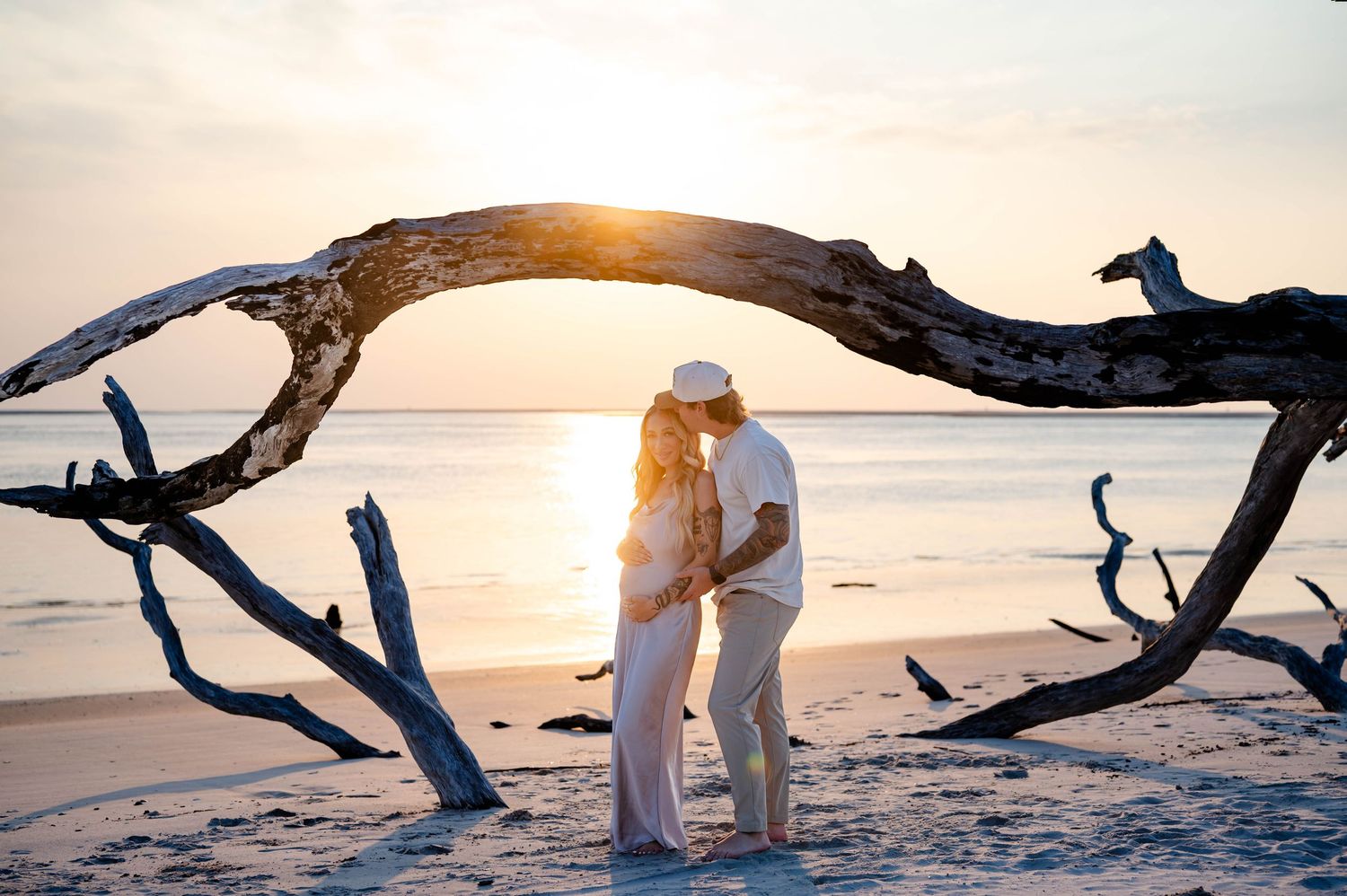 Couple embracing under driftwood arch on beach at sunset.