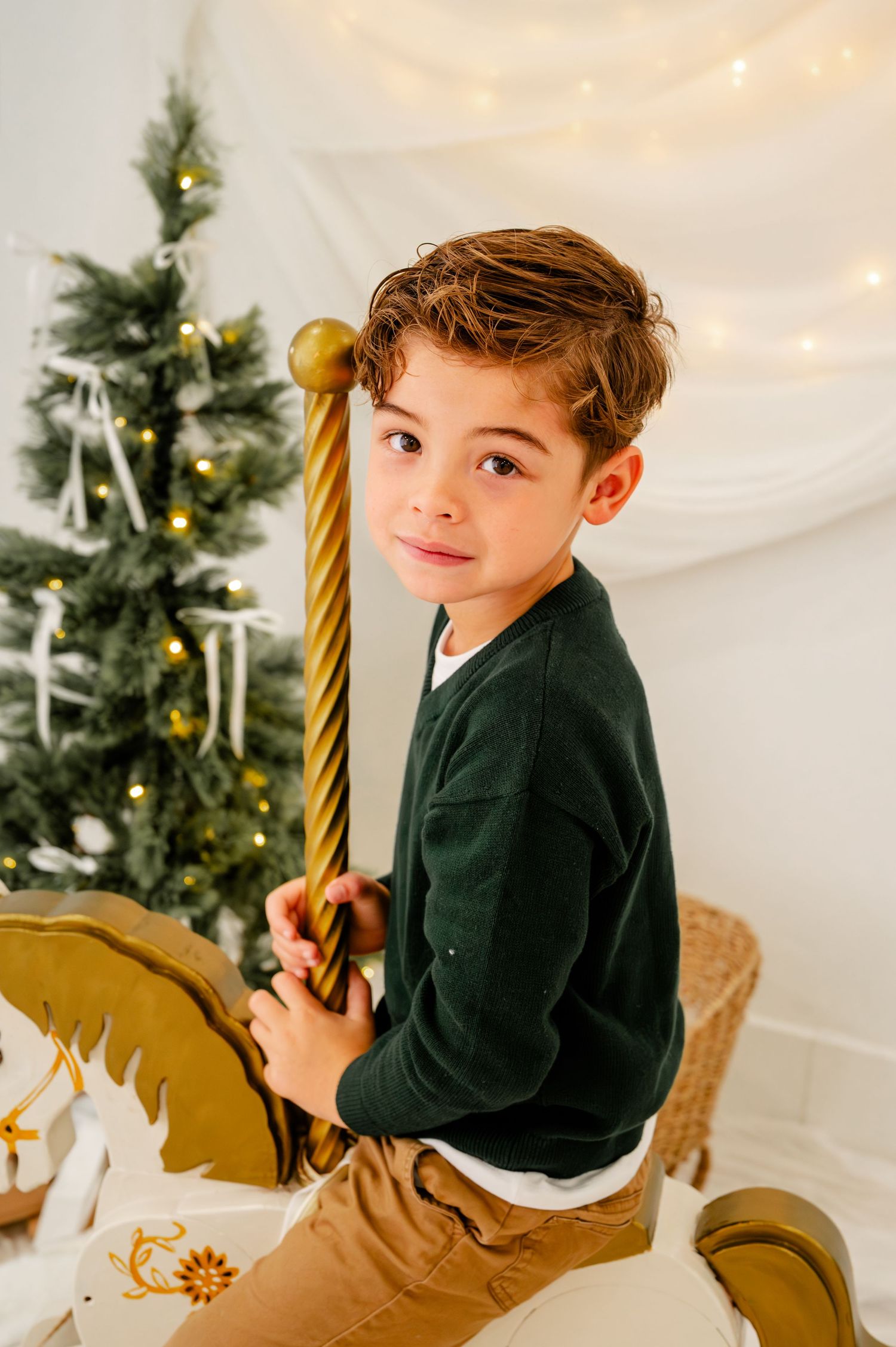 Child in dark blazer poses for holiday portraits with decorated Christmas tree in background.