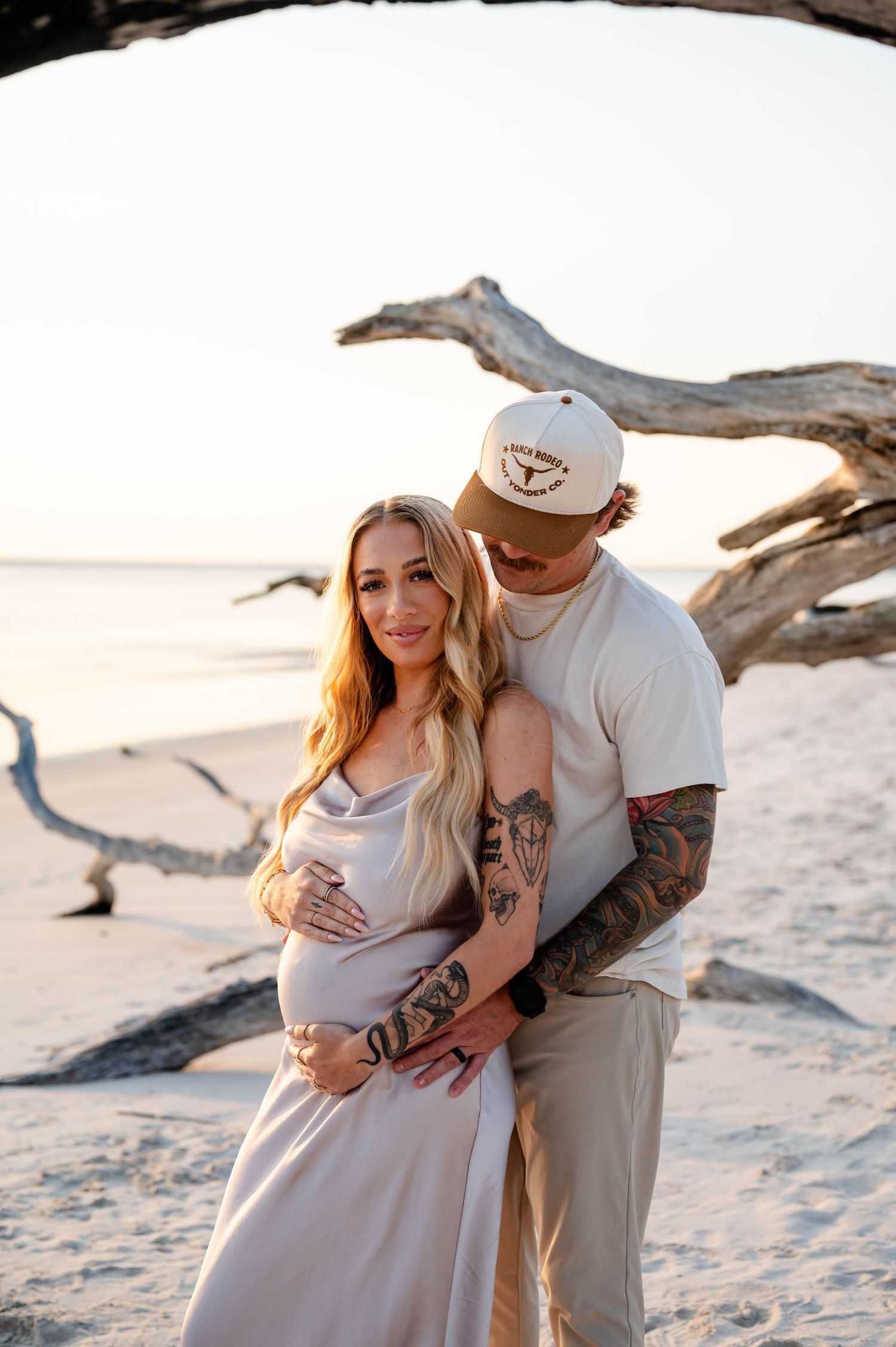 Romantic beach maternity photo with driftwood and sunset backdrop.