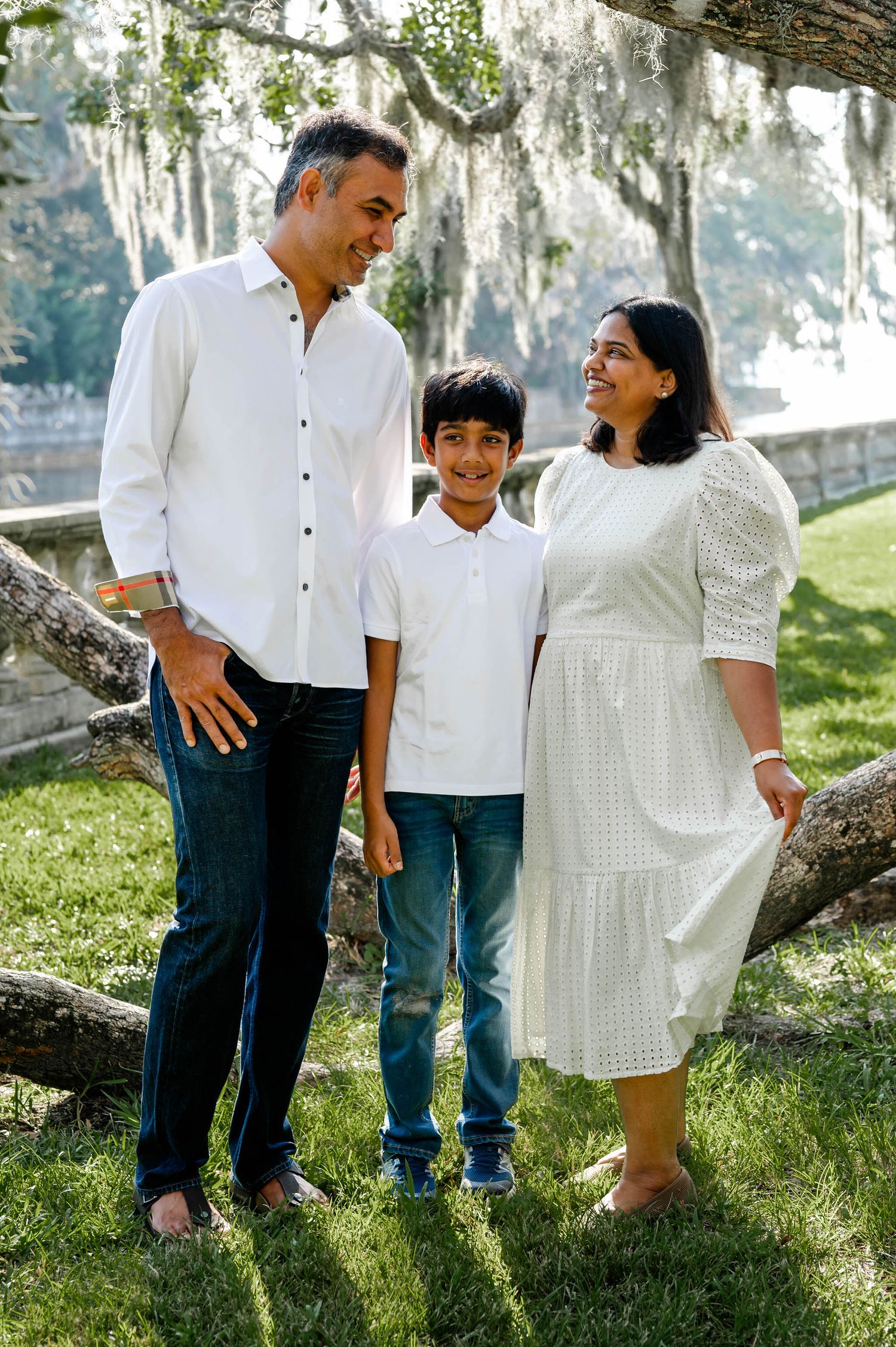 A family wearing white and blue casual outfits poses on green grass near a tree trunk in a park at a spring mini session in jacksonville, fl