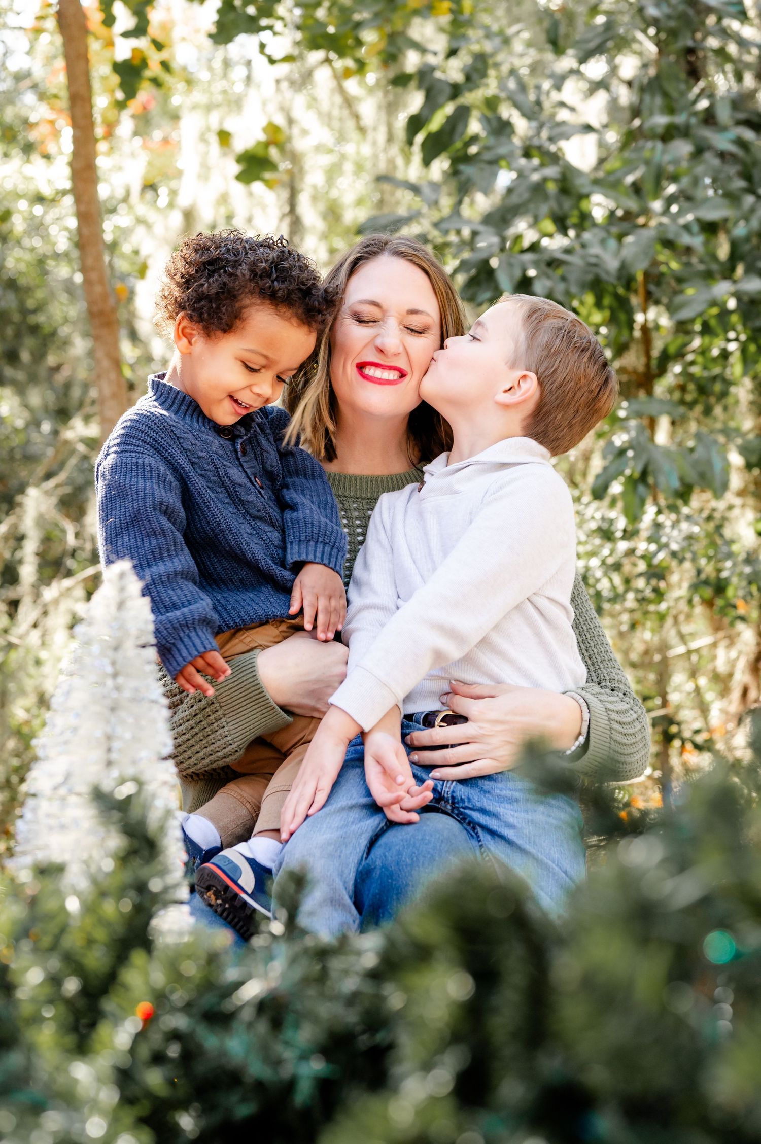 A joyful family photo session in a sunlit wooded area shows tender moments of affection and laughter together.