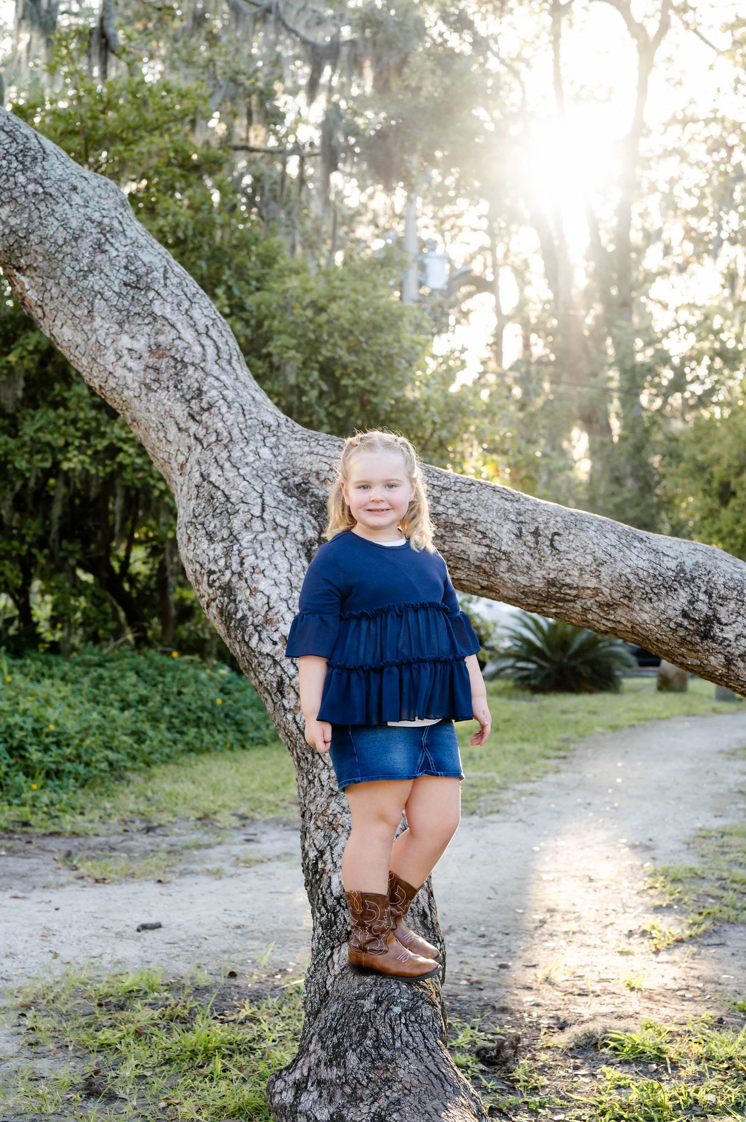A young child in a navy blue dress stands on a curved tree branch in a sunlit wooded area.