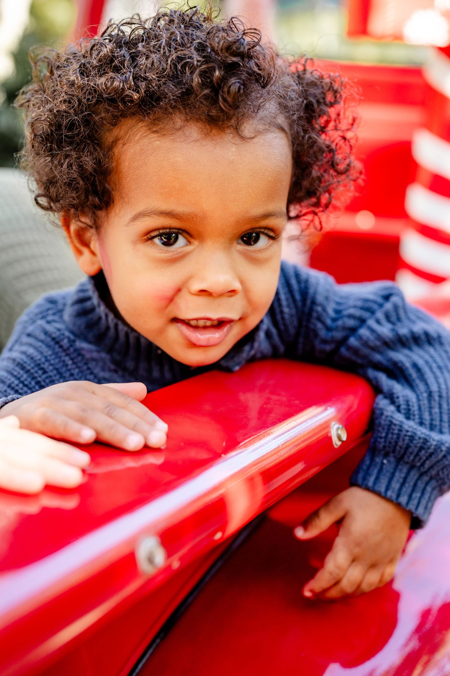 A child in a navy sweater peers over a bright red playground structure with a curious expression.