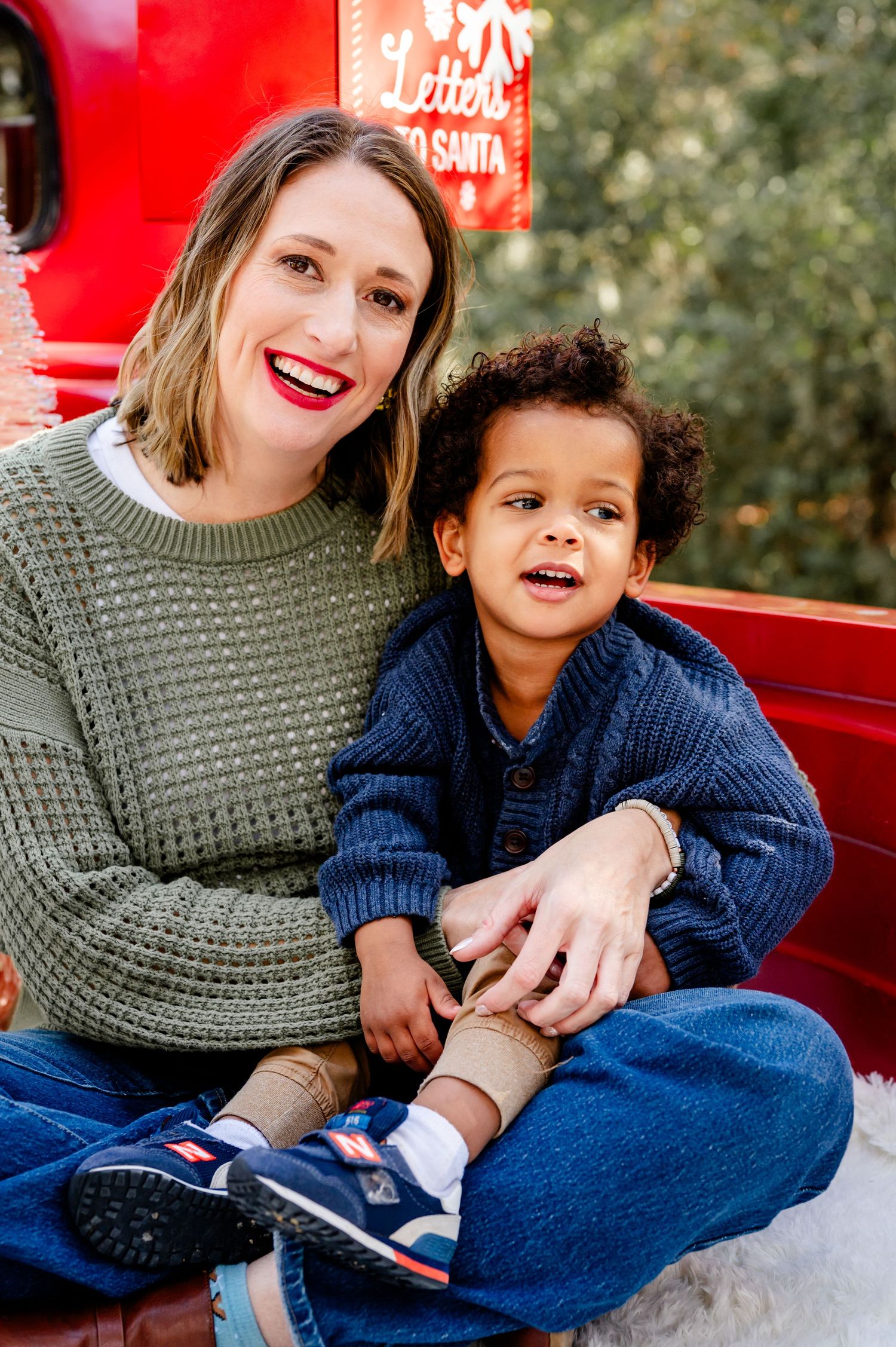 A parent and child sit together on a red surface wearing cozy sweaters and smiling warmly at the camera.