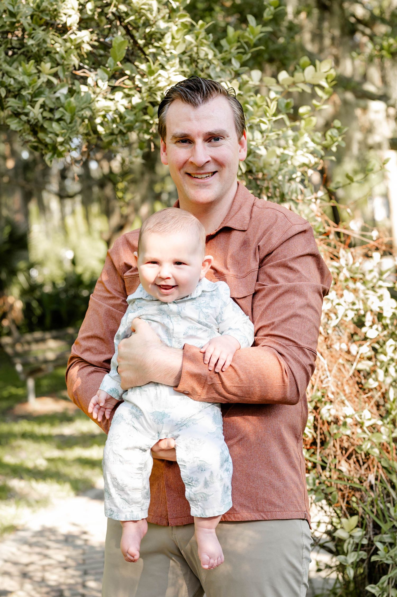 A parent and baby pose together outdoors in a garden setting on a sunny spring day.