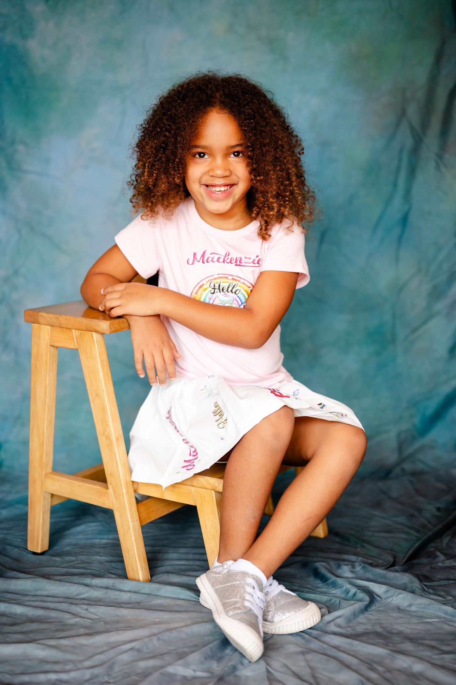 Girl in white outfit sitting on wooden stool against blue backdrop captured by a preschool photographer in Jacksonville FL.