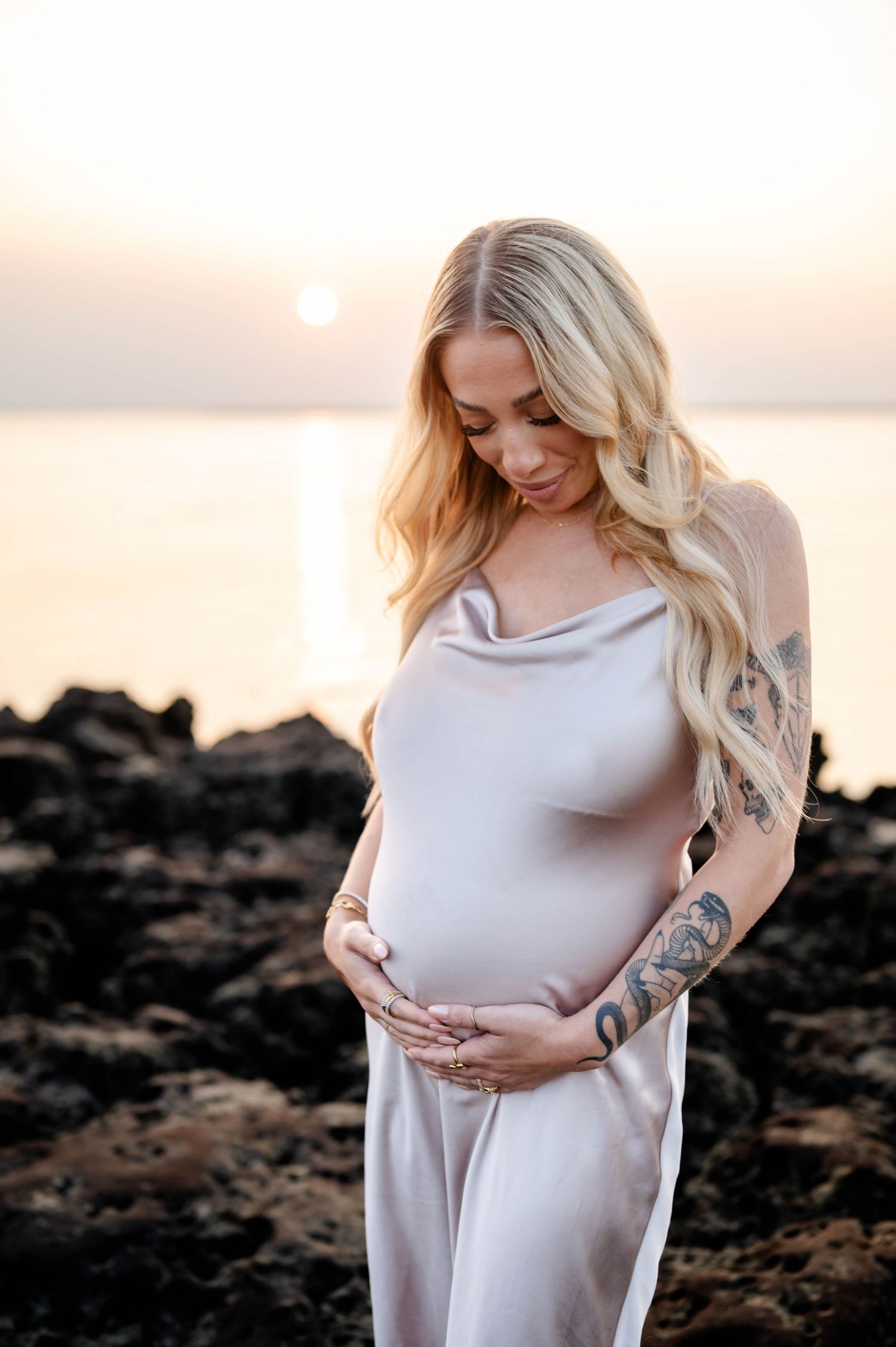 Expectant mother in flowing beige dress poses on rocky beach during golden hour.