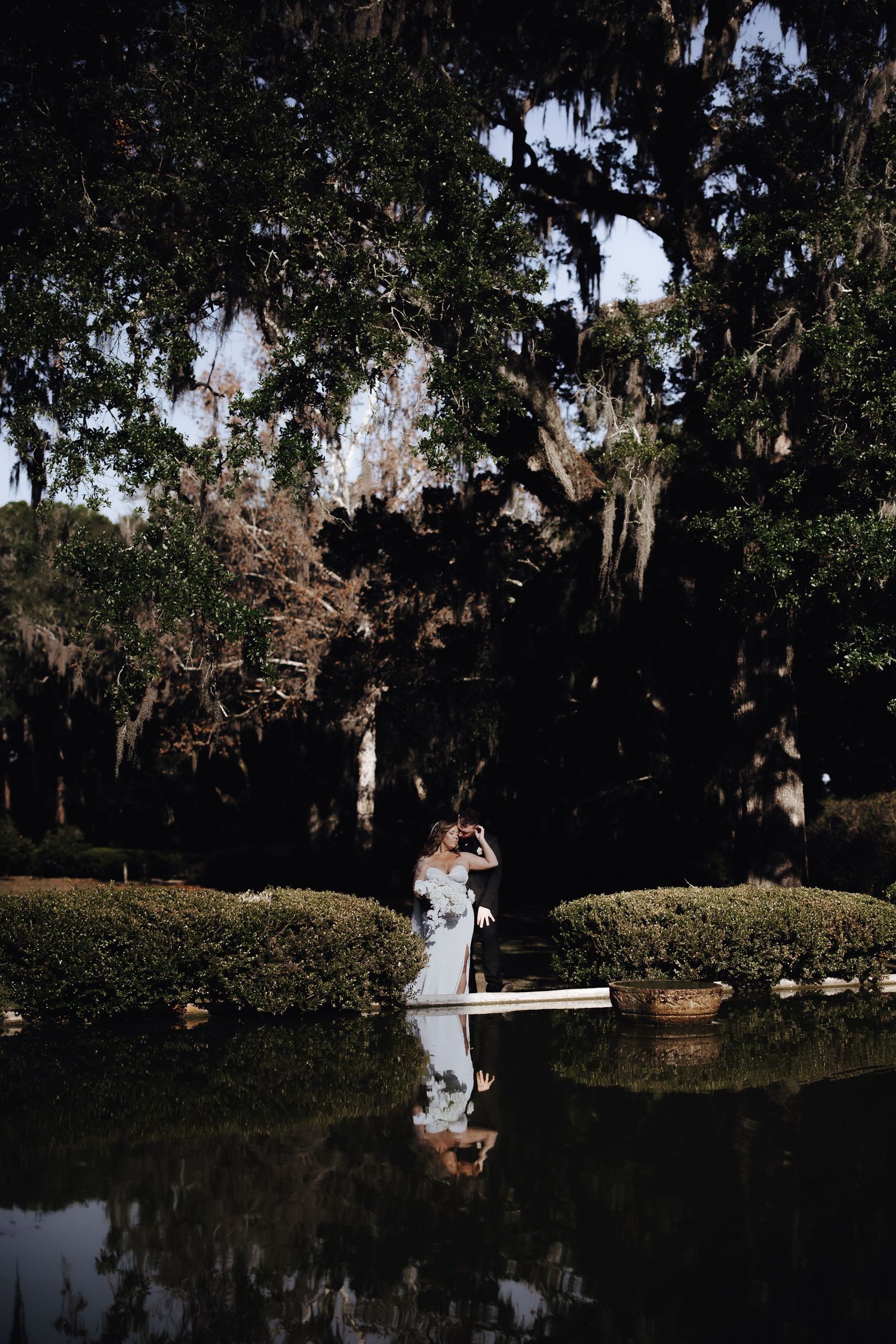 A serene outdoor wedding portrait session at dusk with a couple reflected in a tranquil pond surrounded by Spanish moss trees.