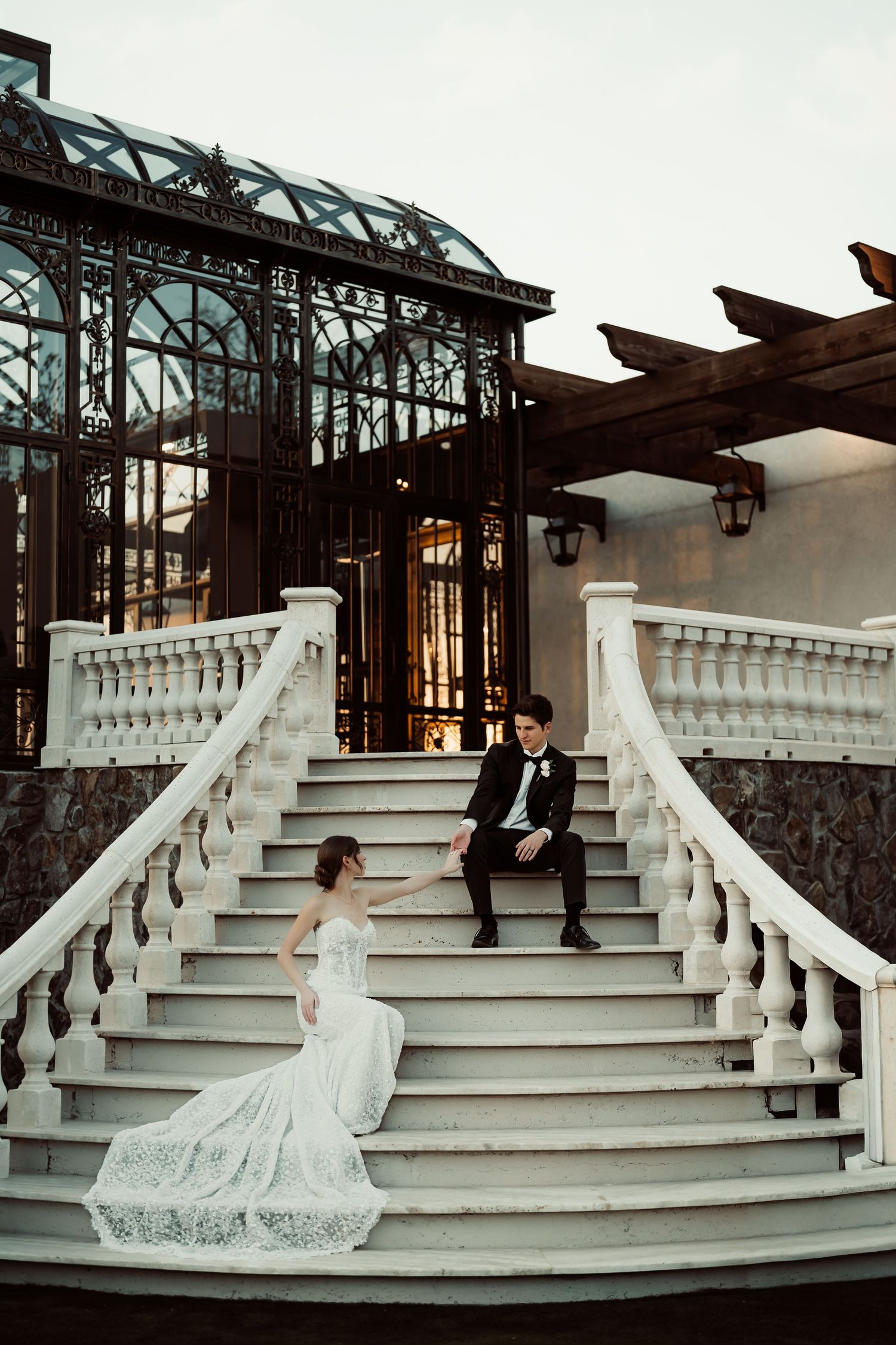 Bride and groom pose on elegant white marble staircase outside a grand venue with ornate iron greenhouse backdrop.