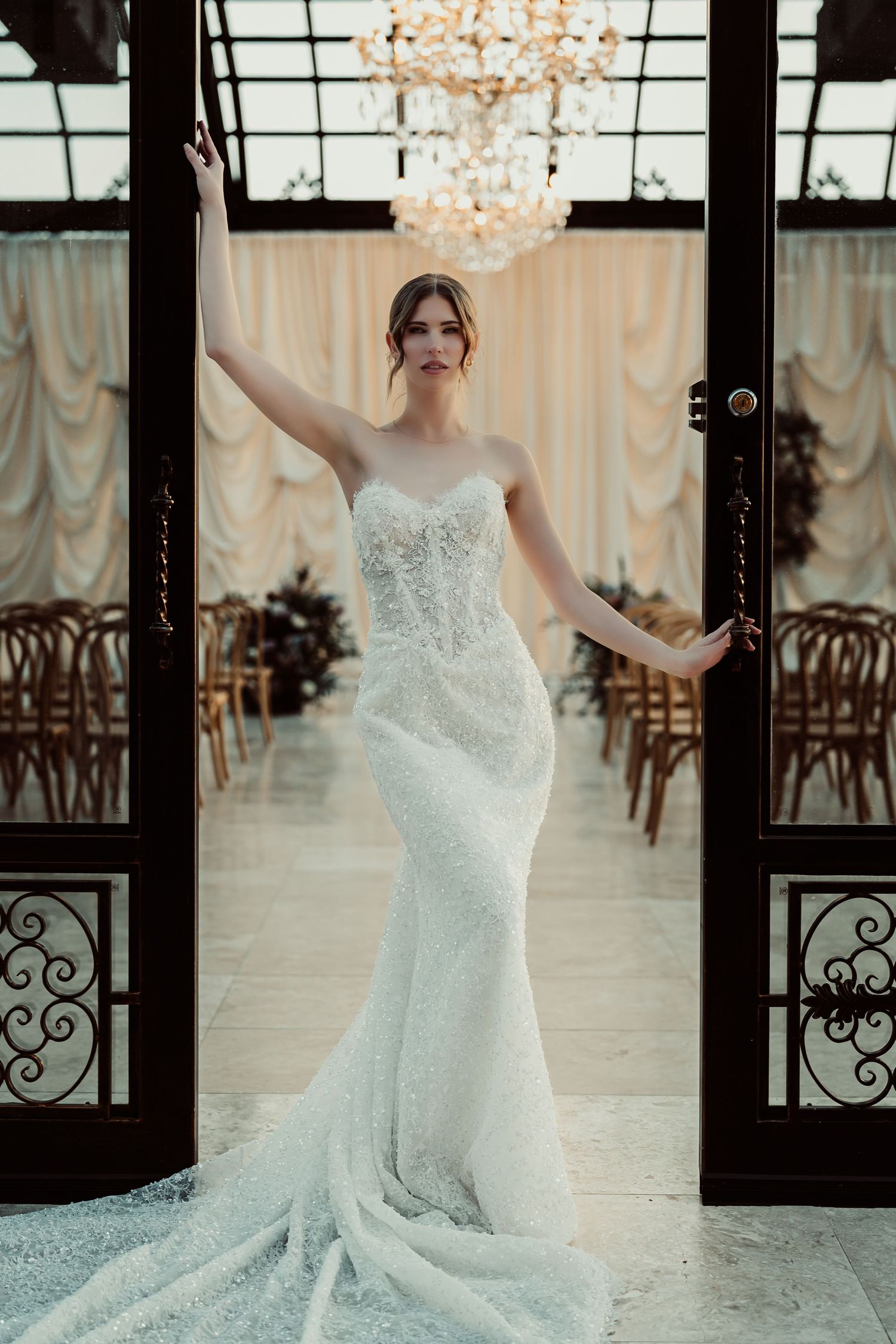 Bride in strapless lace mermaid wedding gown stands in ornate doorway of elegant venue with chandelier.