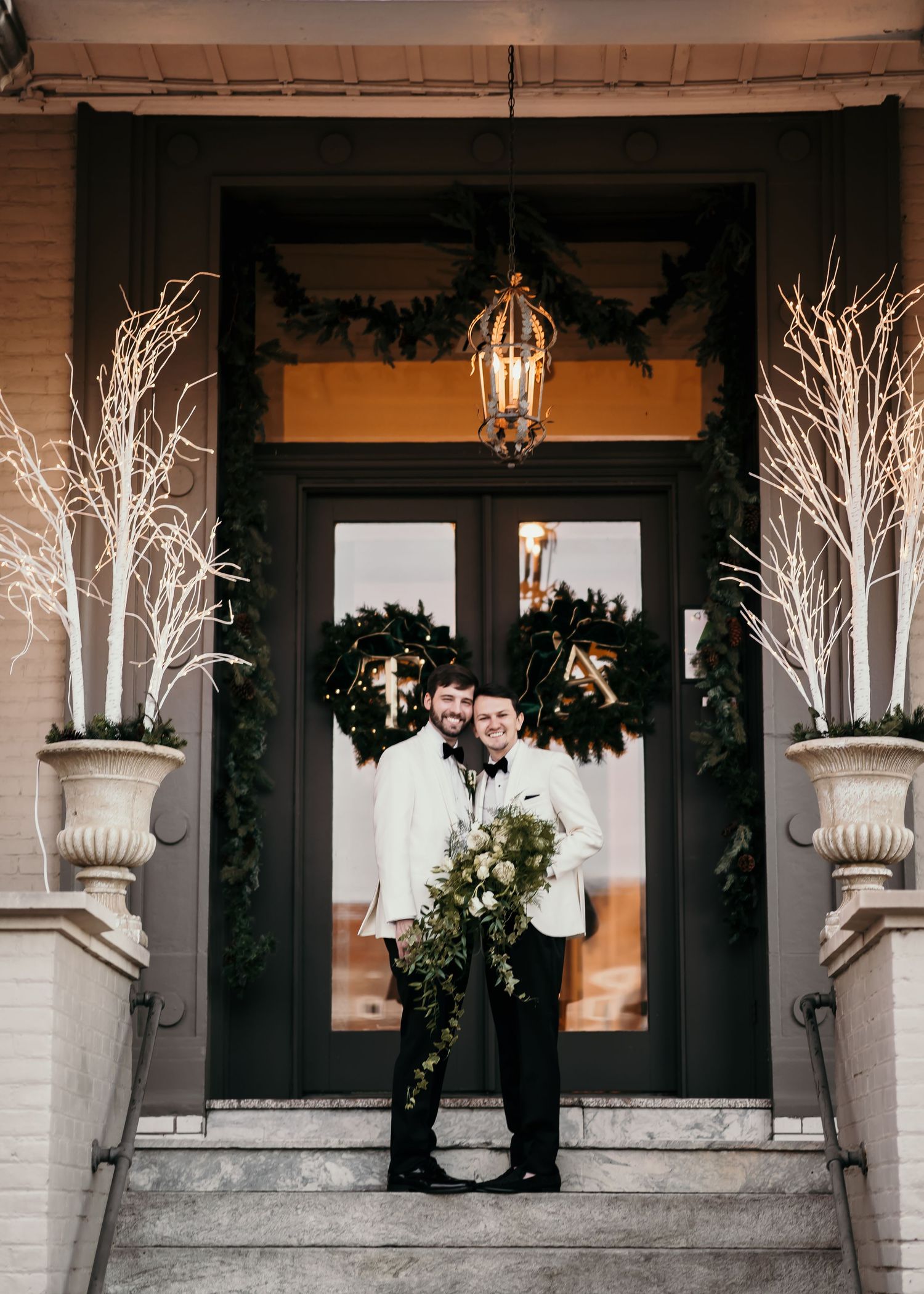 A festive winter porch decorated with white branches, lanterns and evergreen wreaths on dark doors.