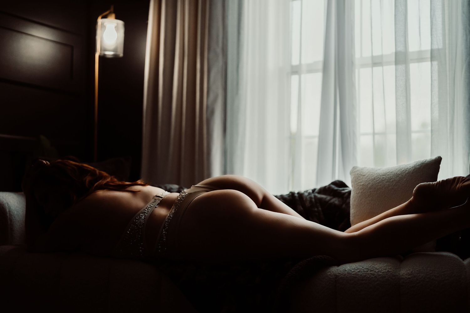 A woman in lingerie reclines on a bed in a dimly lit room with soft light filtering through sheer curtains.