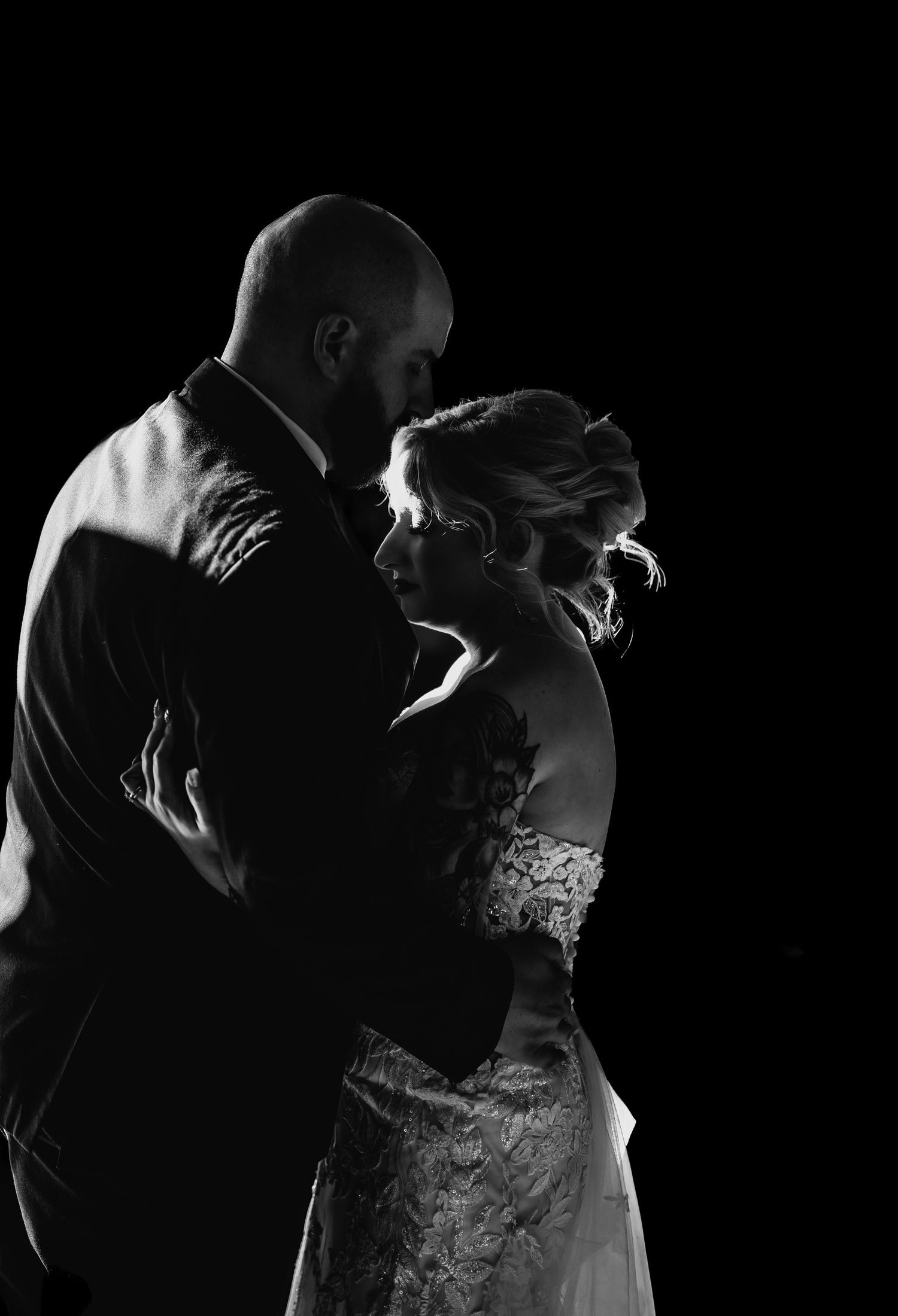Black and white wedding photo of bride and groom sharing an intimate moment against a dark background.