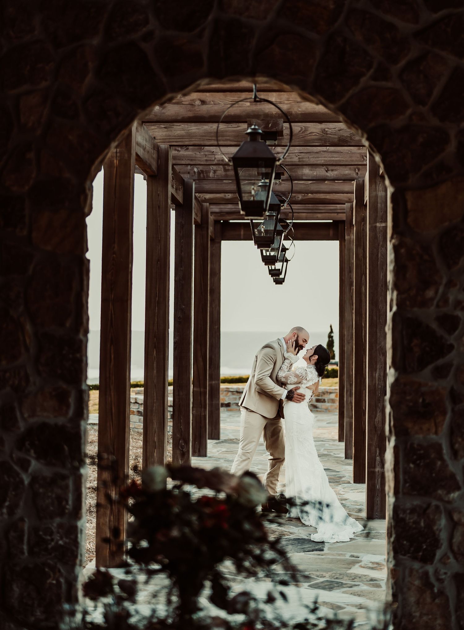 A bride and groom share a kiss under a rustic stone archway with hanging lanterns, ocean view in background.