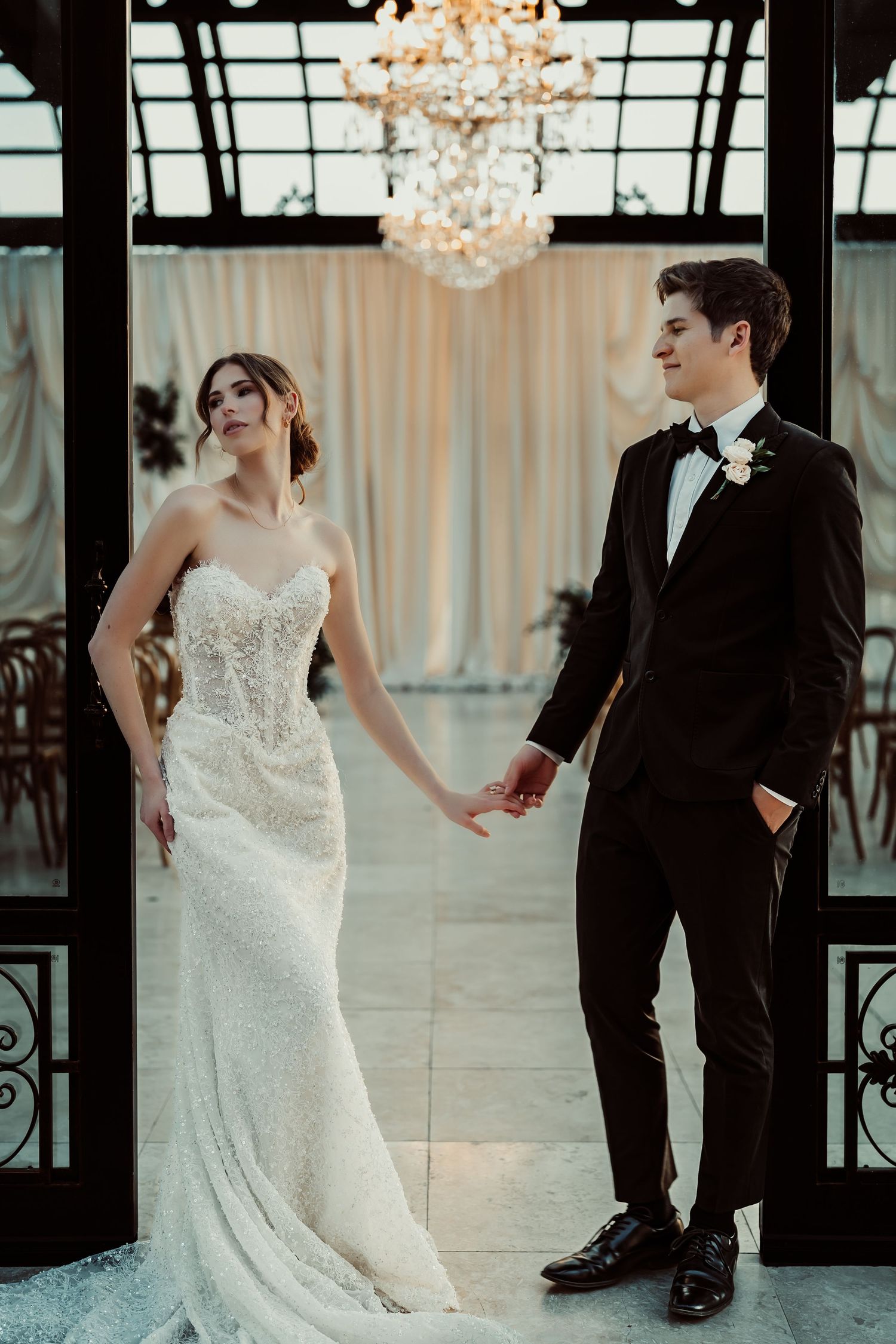 Elegant bride in lace gown and groom in black tuxedo holding hands under a crystal chandelier at a wedding venue.