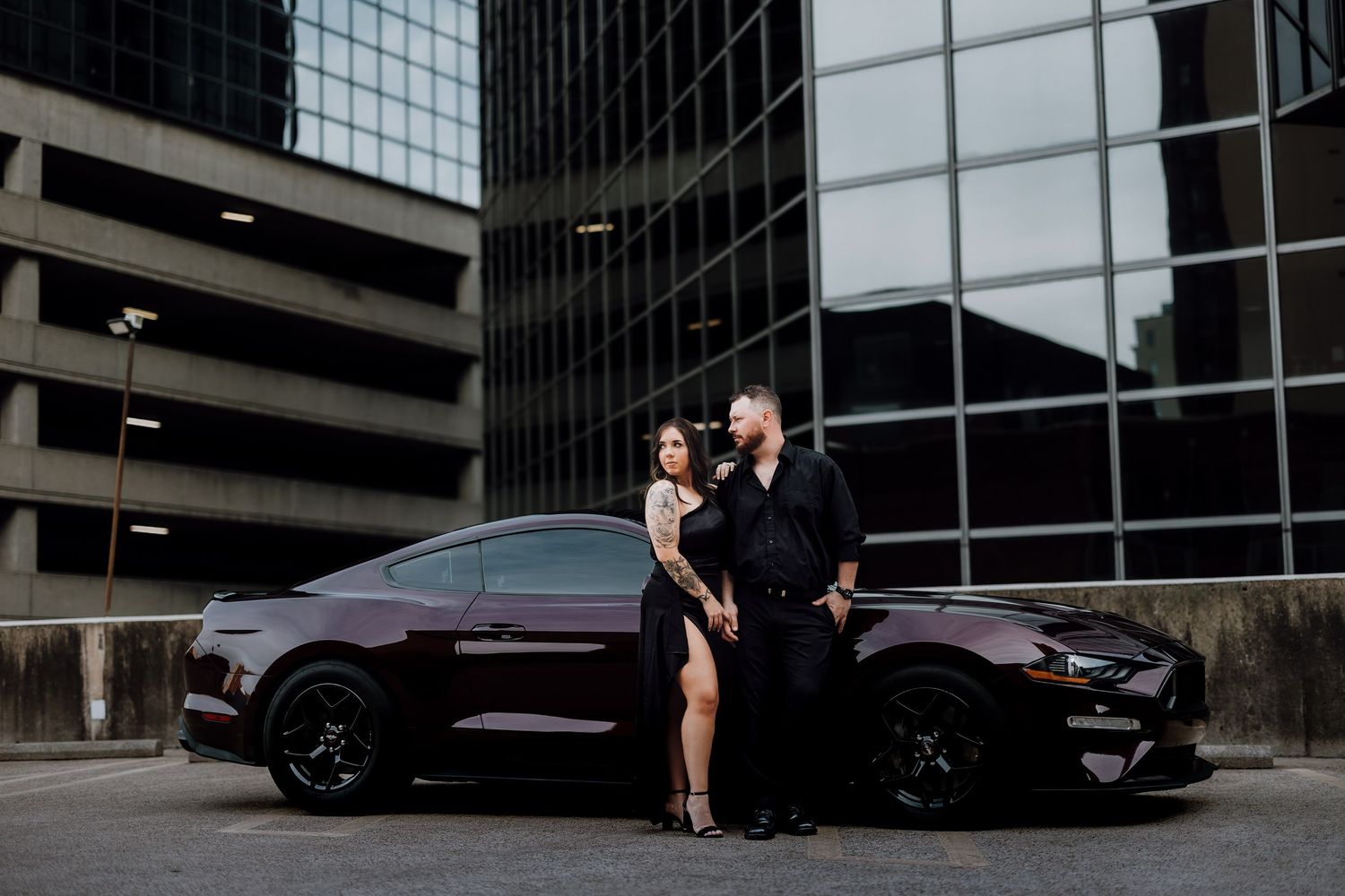 Couple posing beside a dark maroon Ford Mustang in front of modern glass buildings for an engagement photo.