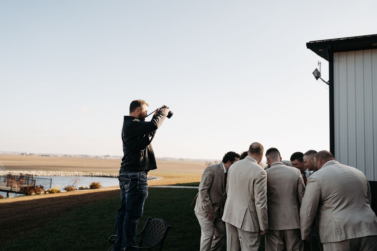 A group of wedding guests stand in formal attire at an outdoor ceremony against a rural landscape backdrop.