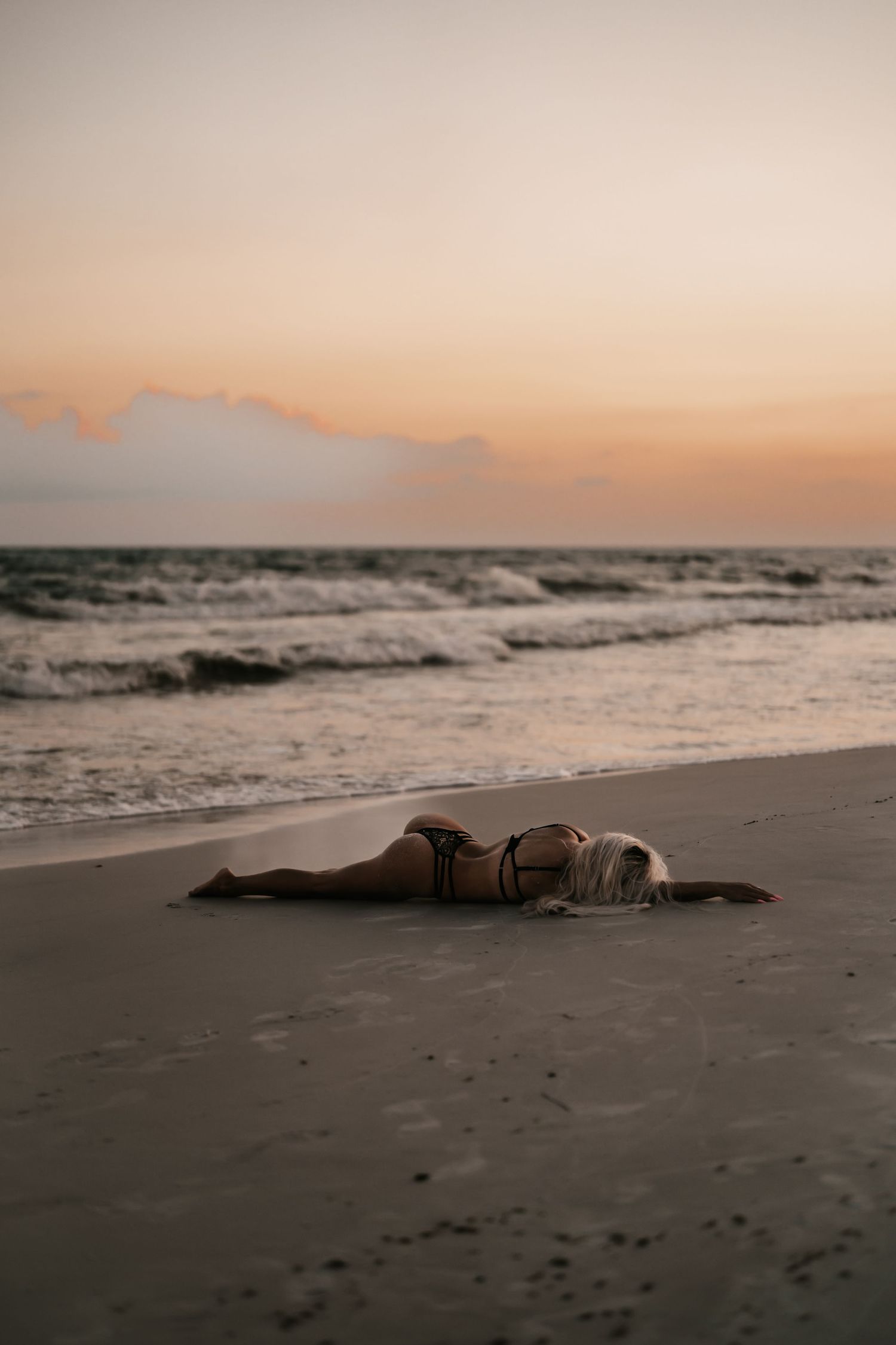 A woman in a bikini lies on a sandy beach at sunset with gentle ocean waves in the background.