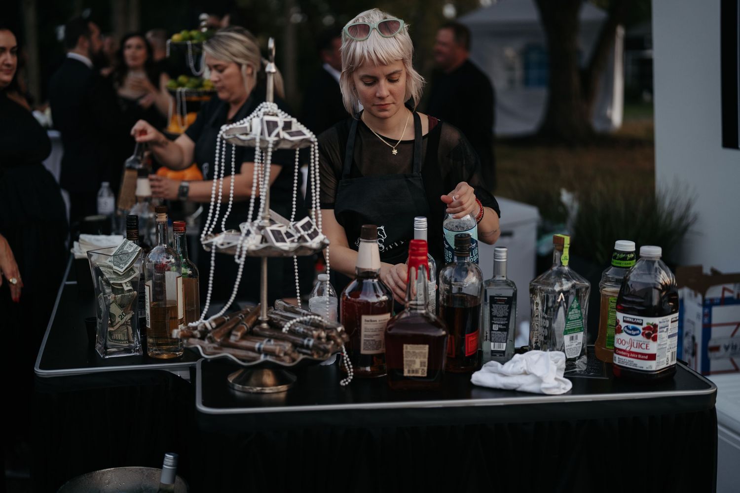 Various liquor bottles and cocktail glasses line up on a dark bar counter in a dimly lit establishment.