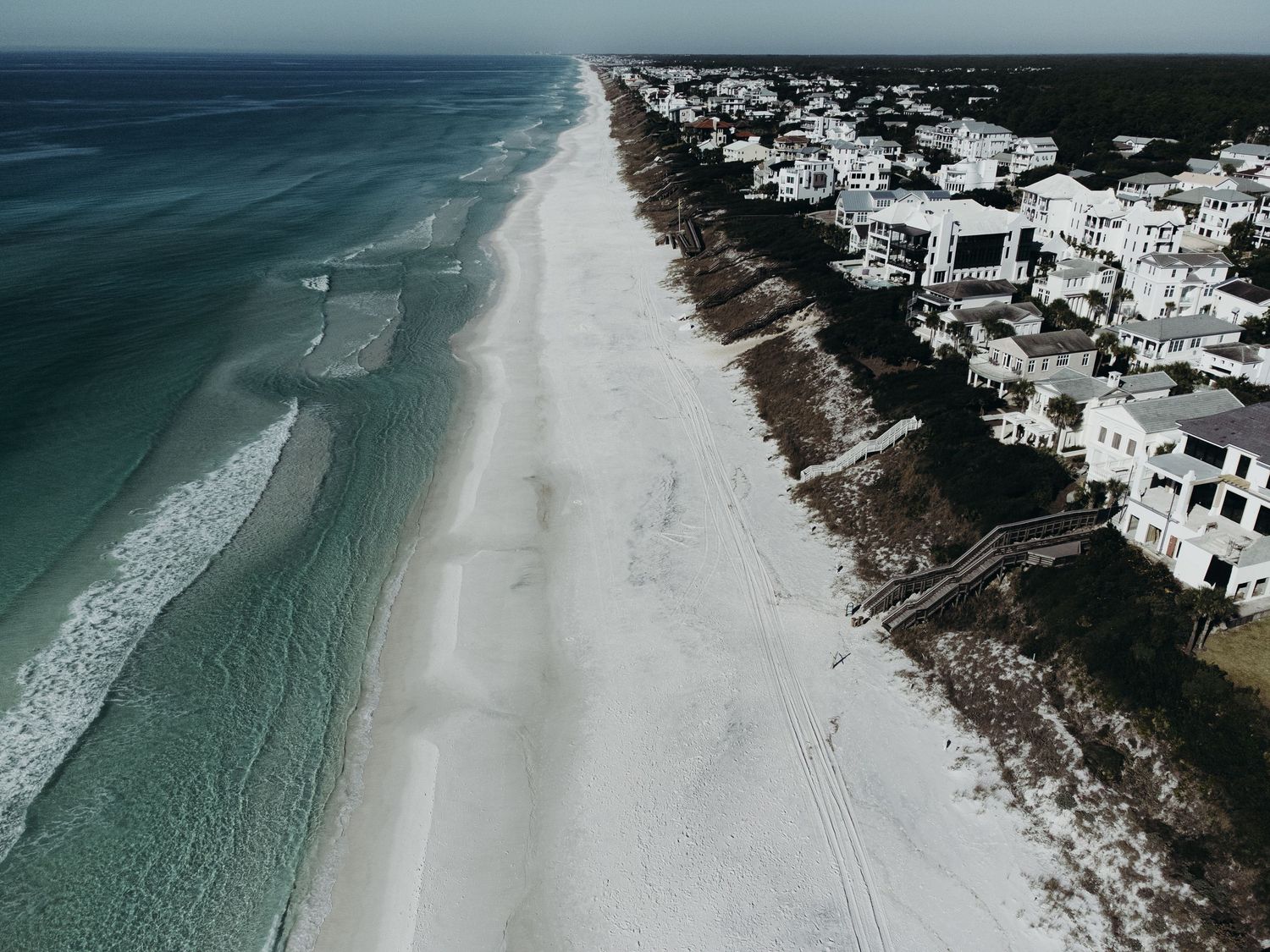 Aerial view of white sandy beach with turquoise waves beside luxury coastal homes and lush trees under overcast skies.