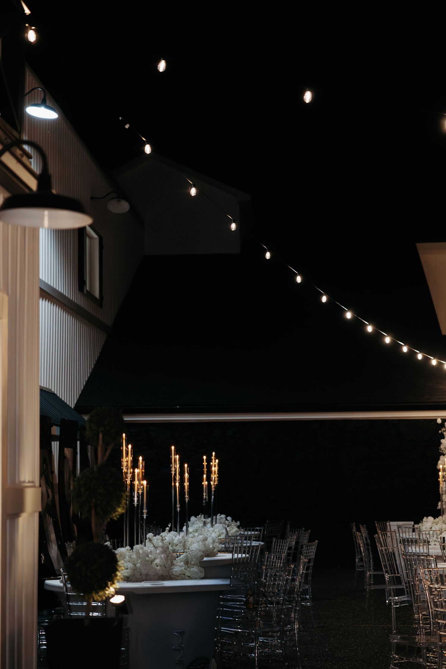 String lights illuminate a dark event space with clear chairs and white decor for an elegant reception setup.