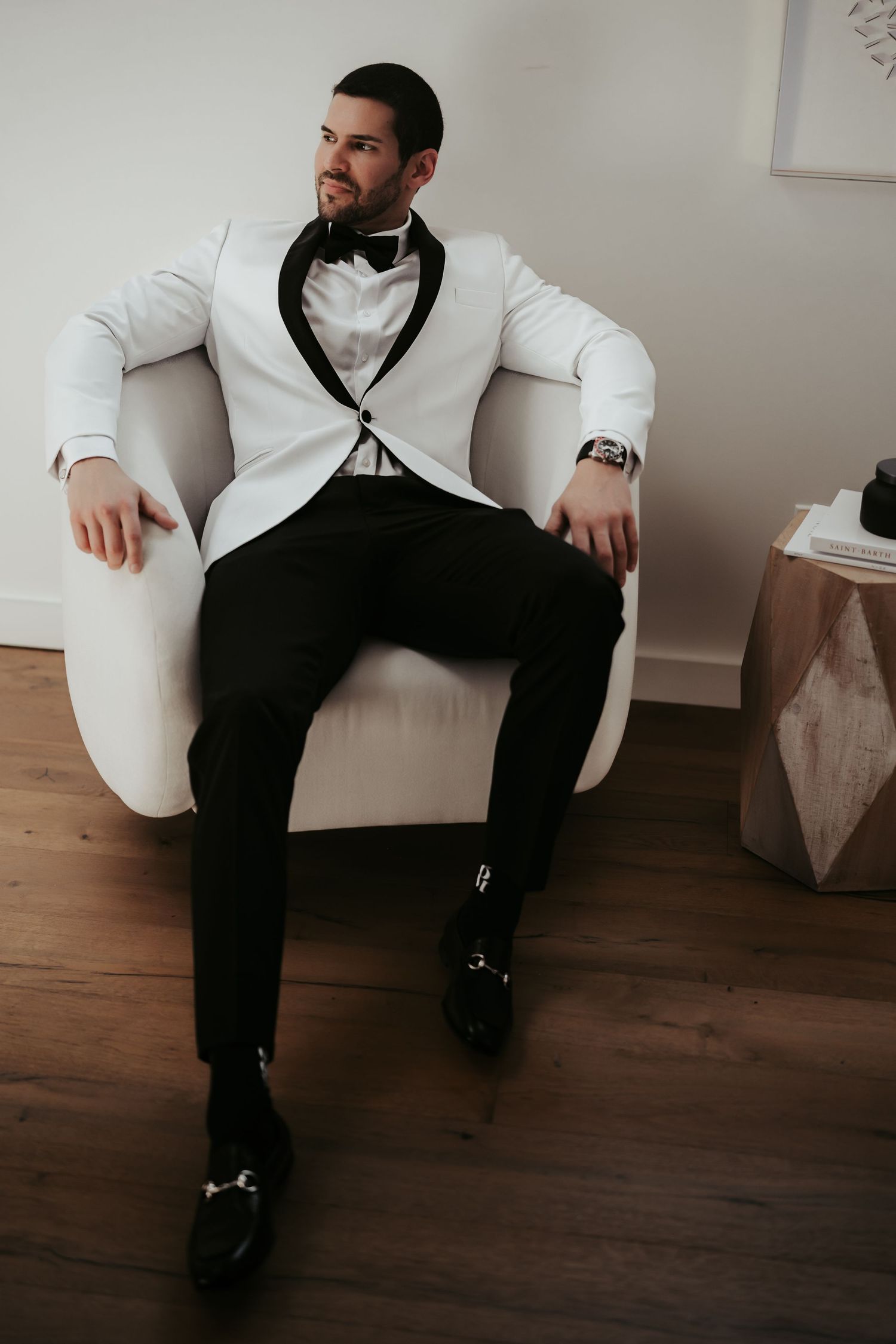 Elegant groom in white tuxedo jacket and black bow tie lounges in modern white armchair.