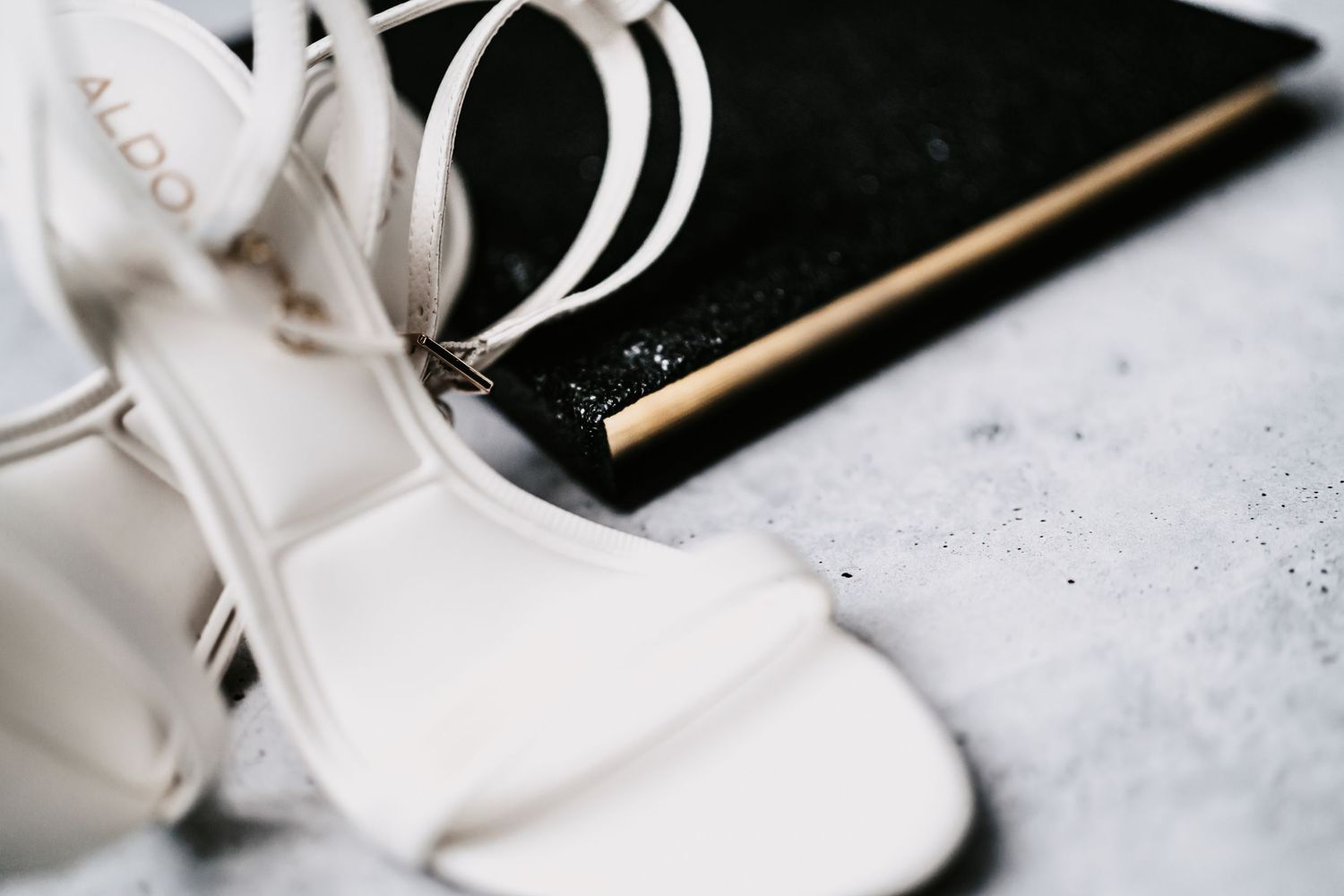 White strappy ALDO heeled sandals beside a black and gold sequined clutch purse on a marble surface.