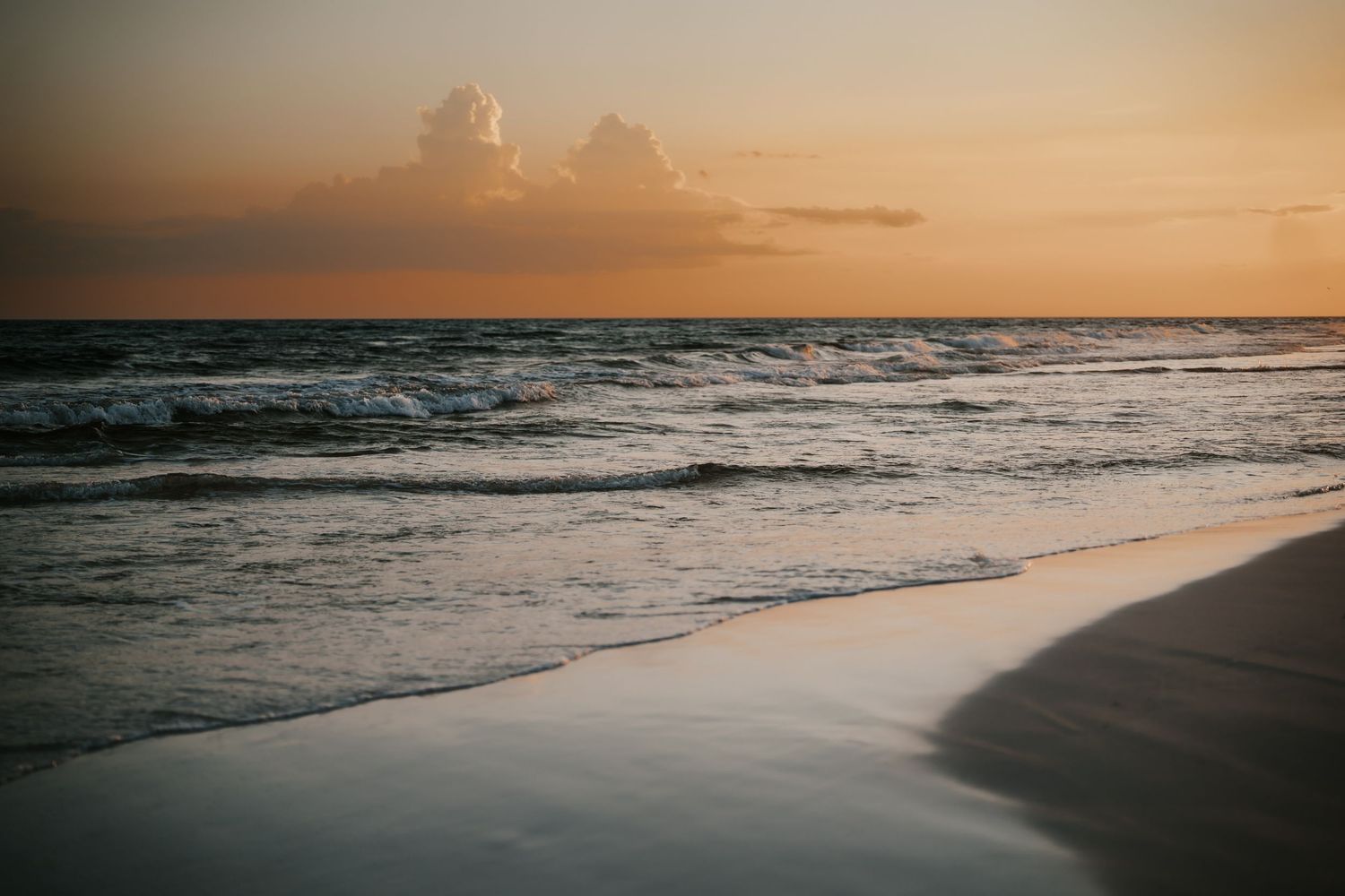 Golden sunset over calm ocean waves gently washing onto a sandy beach with dramatic clouds on the horizon.