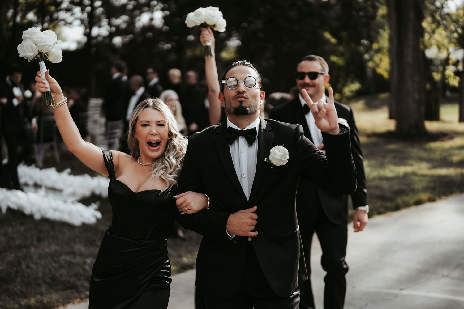 Joyful wedding party in black formal attire celebrating outdoors, bride raising bouquet, groom in tuxedo with boutonniere.