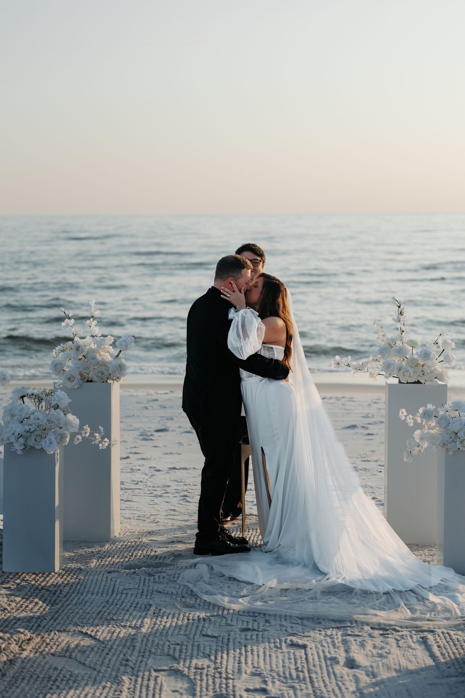 A romantic beach wedding ceremony at sunset with white floral arrangements and a couple sharing an intimate moment.
