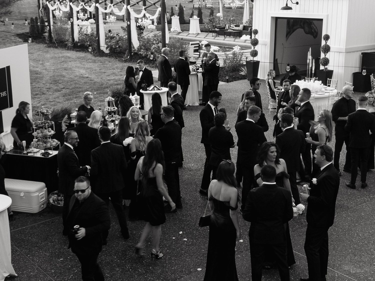 A black and white photograph shows a formal outdoor cocktail reception with guests mingling in elegant attire.