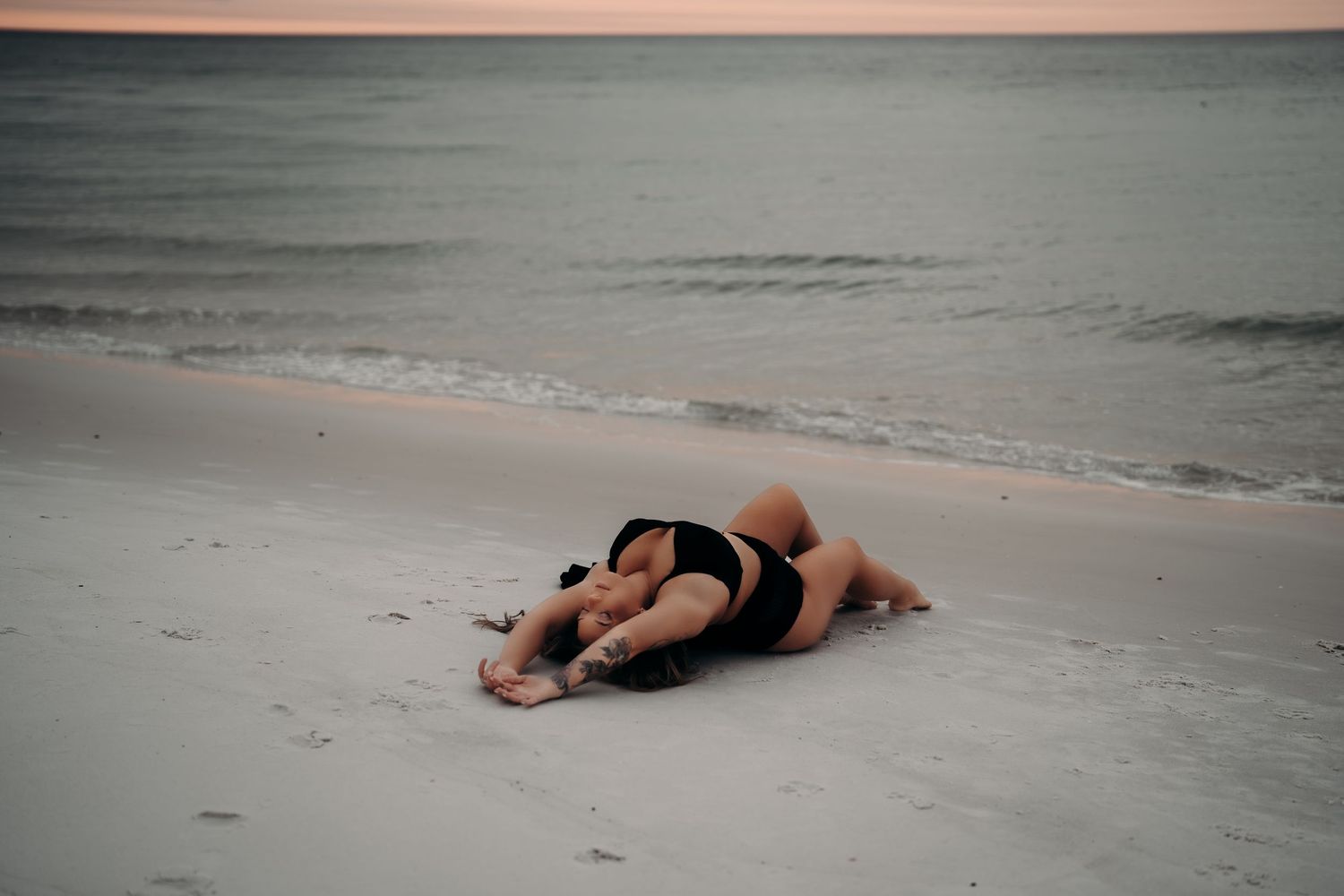 Two people embracing on a sandy beach at dusk, lying together near the ocean shoreline in black swimwear.