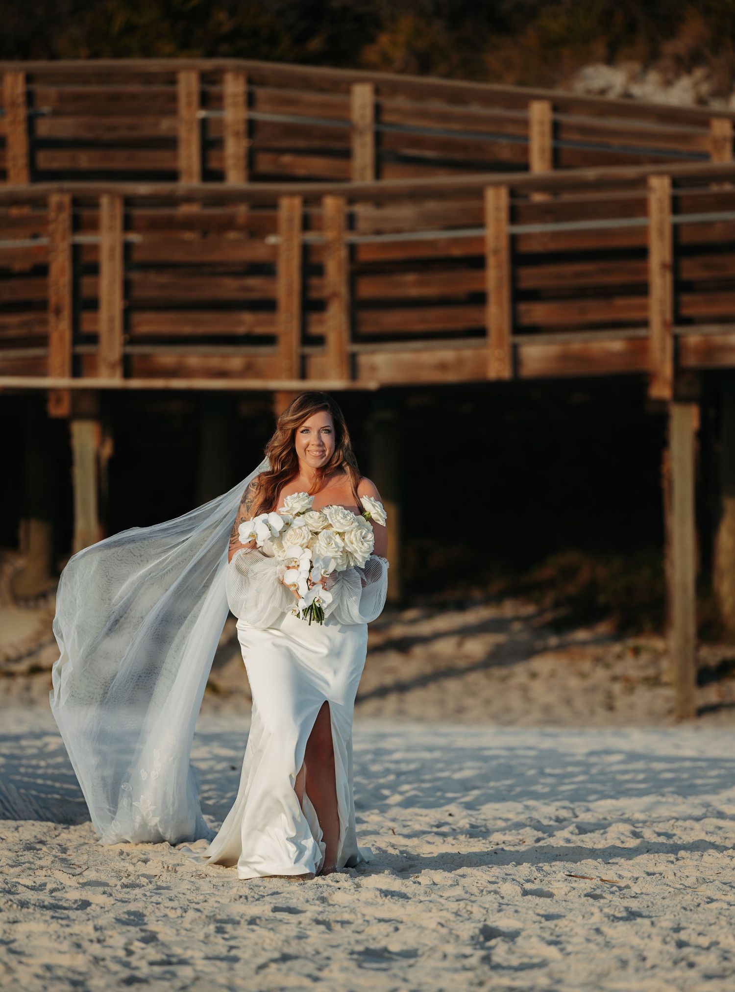 A bride in a flowing white dress with a long veil poses on a beach with a wooden boardwalk in the background.