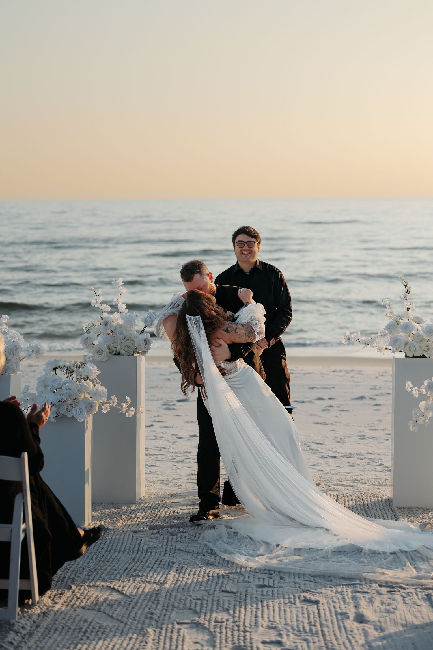 A beachside wedding ceremony with white floral pedestals and a couple embracing during golden hour.