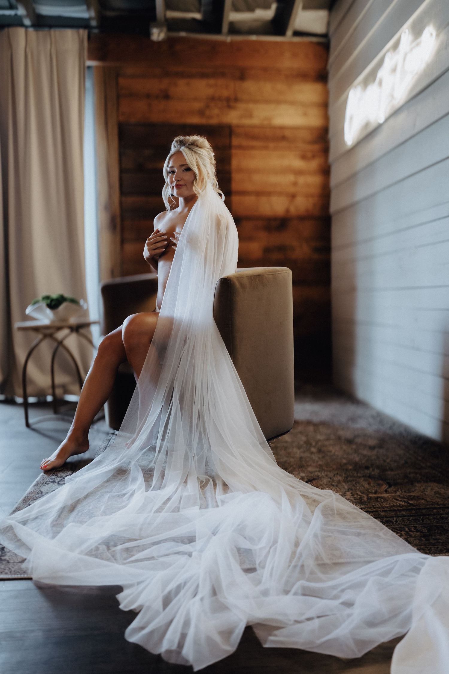 Bride in sheer white tulle robe seated elegantly in a rustic room with wood-paneled walls during bridal boudoir session.