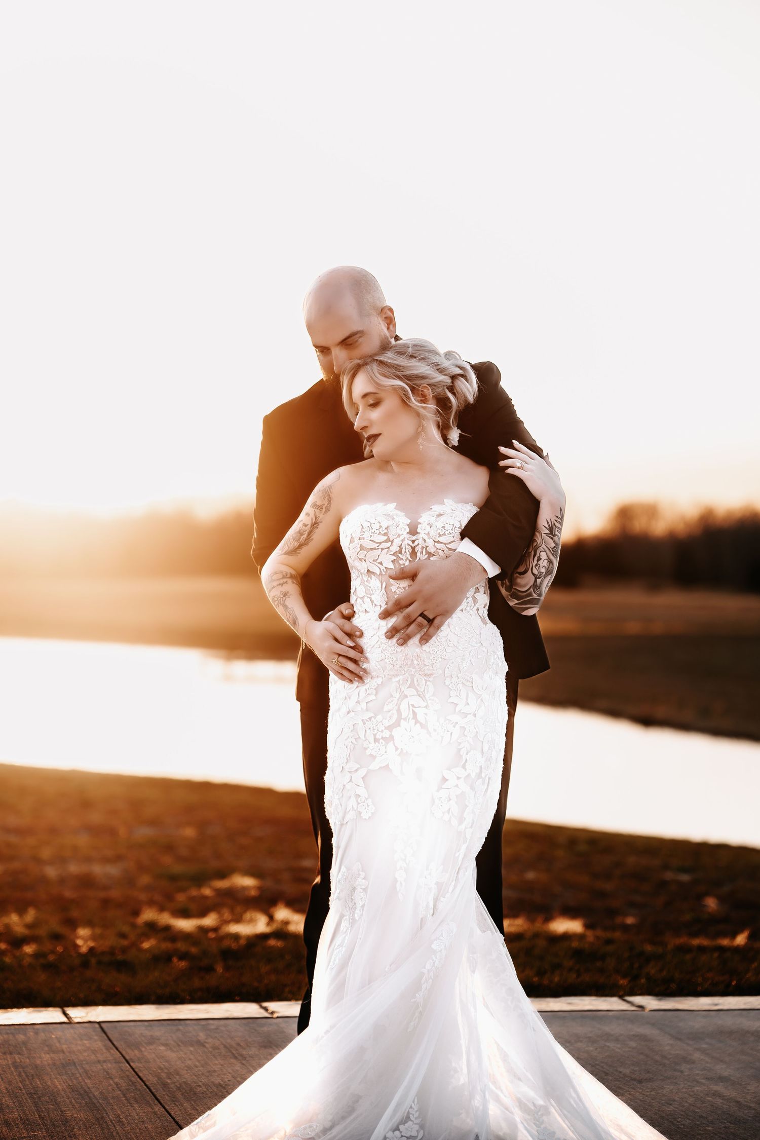 Bride in lace mermaid gown embraced by groom in black suit during golden sunset by a serene lake.