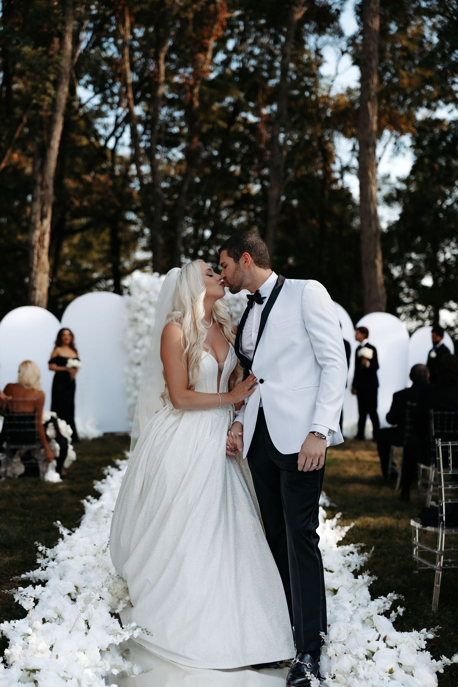 Bride and groom share a kiss at outdoor wedding ceremony with white floral aisle and balloon arch backdrop.