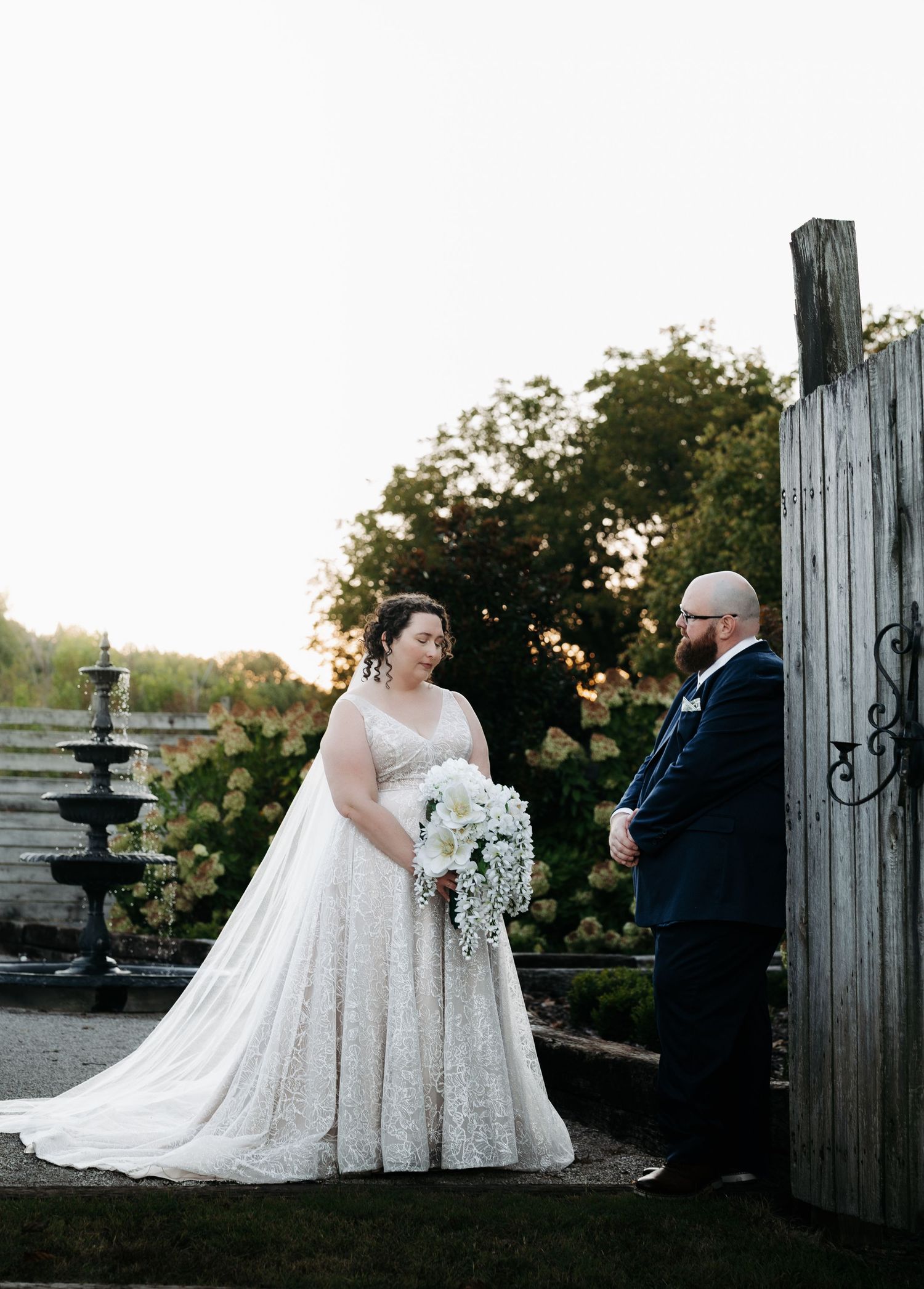 Wedding couple poses by rustic wooden fence and fountain during golden hour sunset.
