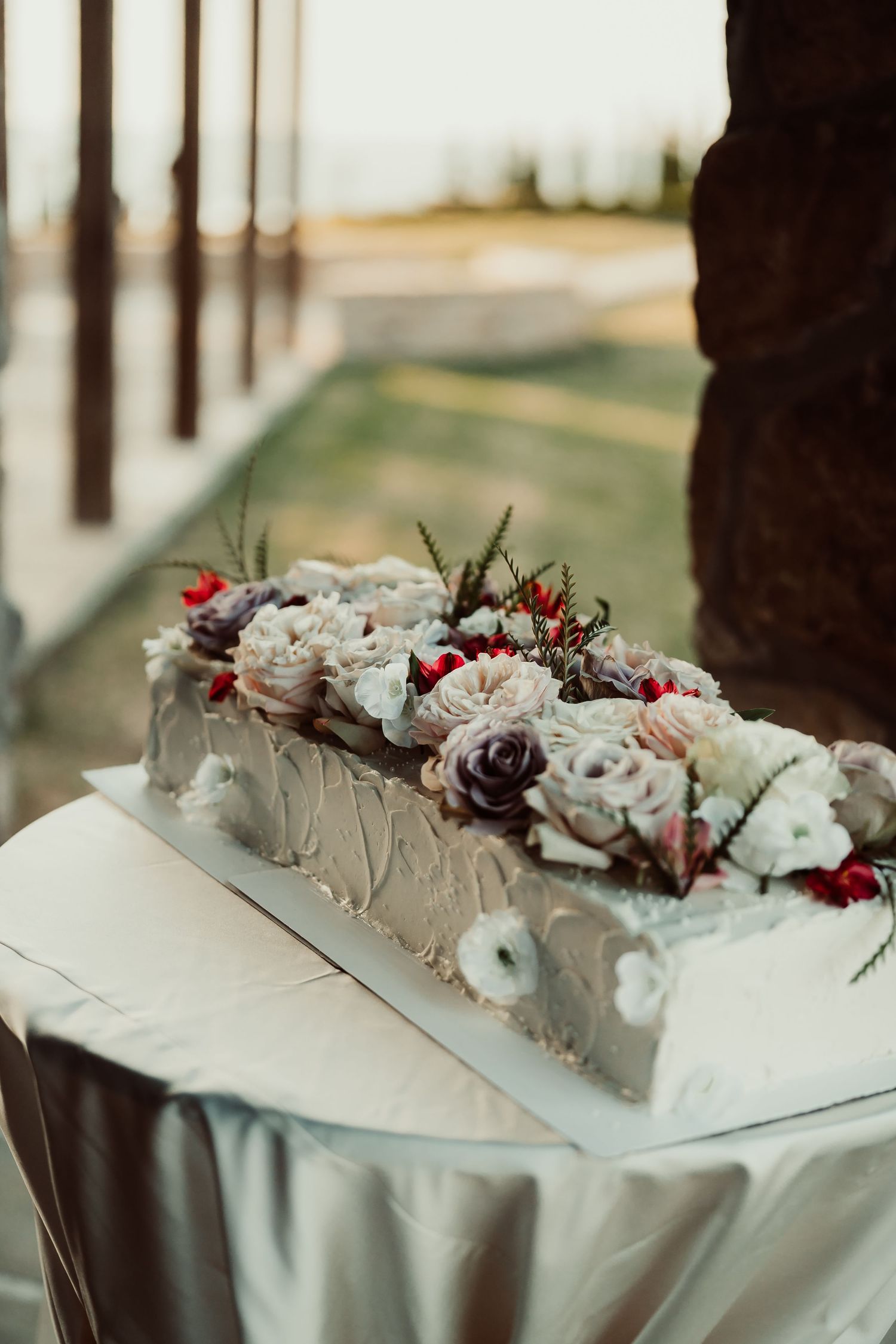 Elegant rectangular wedding cake topped with pastel floral decorations on a white draped table outdoors.