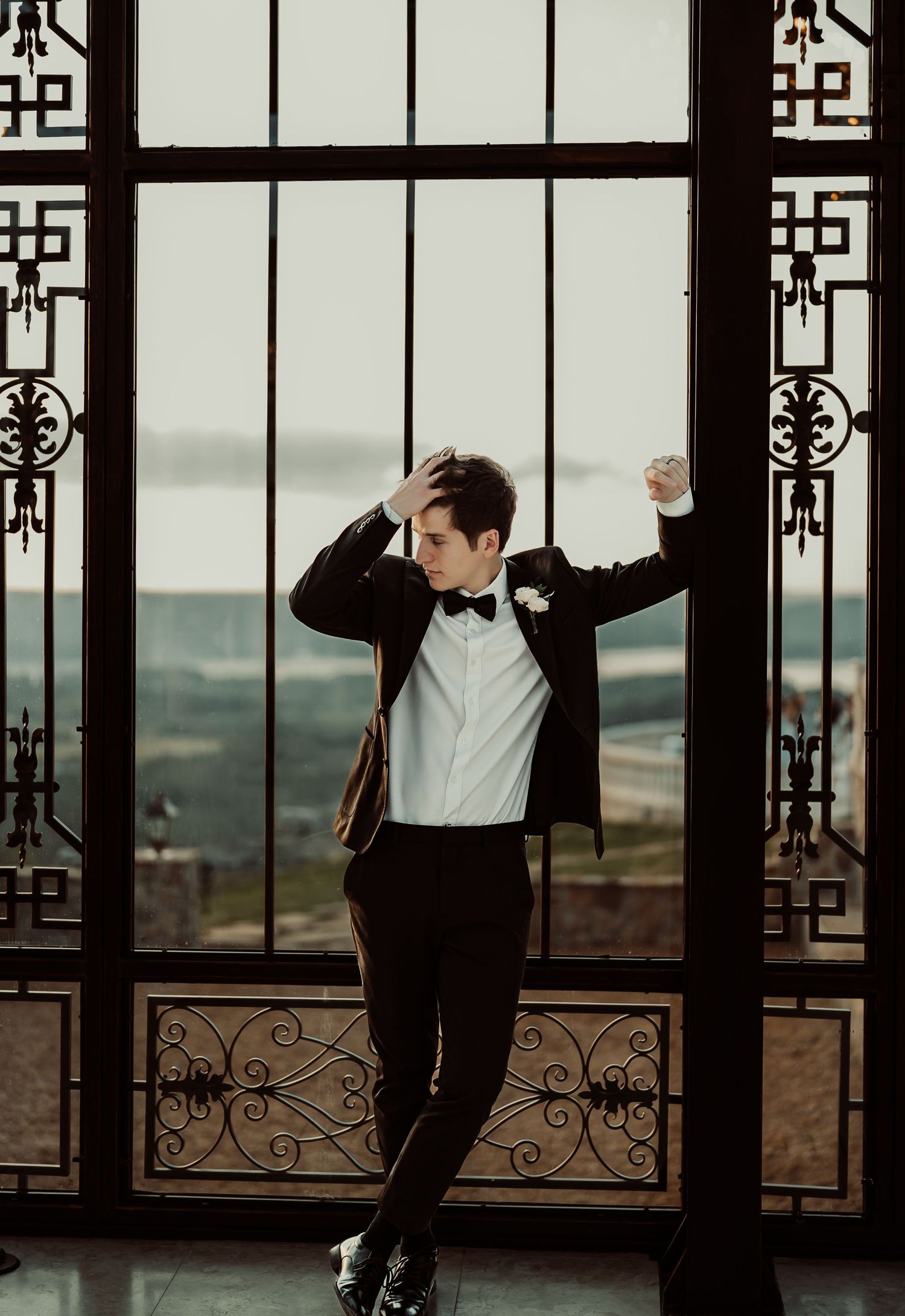 Groom in black tuxedo and bow tie poses by ornate iron gates with scenic countryside view.