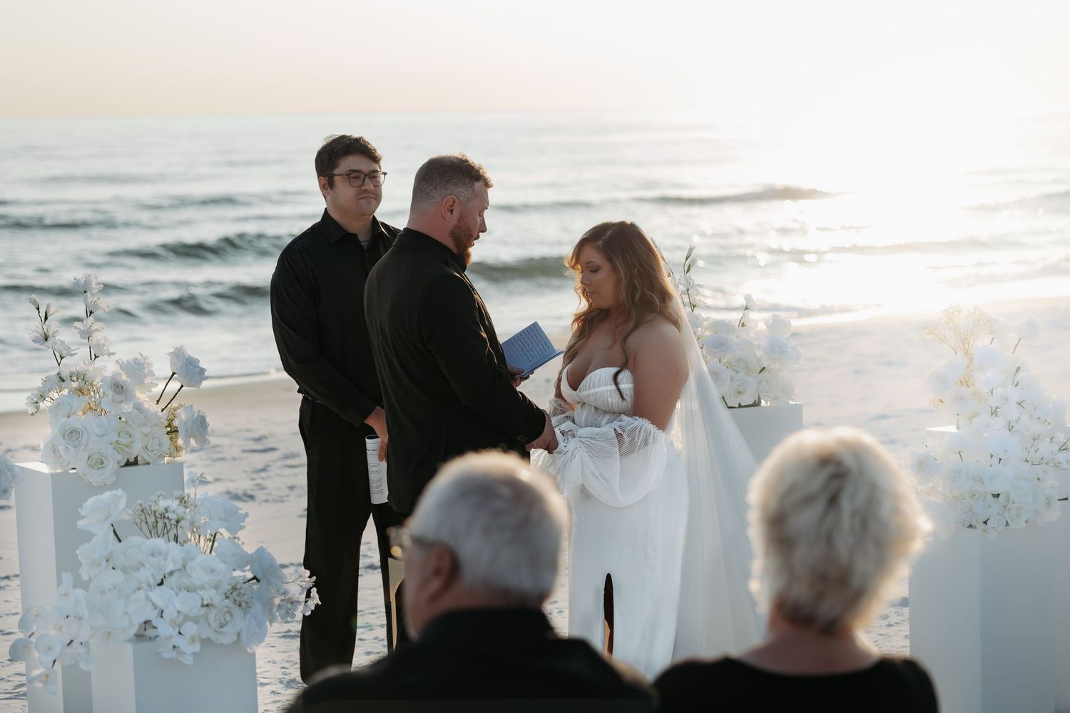 A romantic beach wedding ceremony at sunset with people gathered near the water's edge.