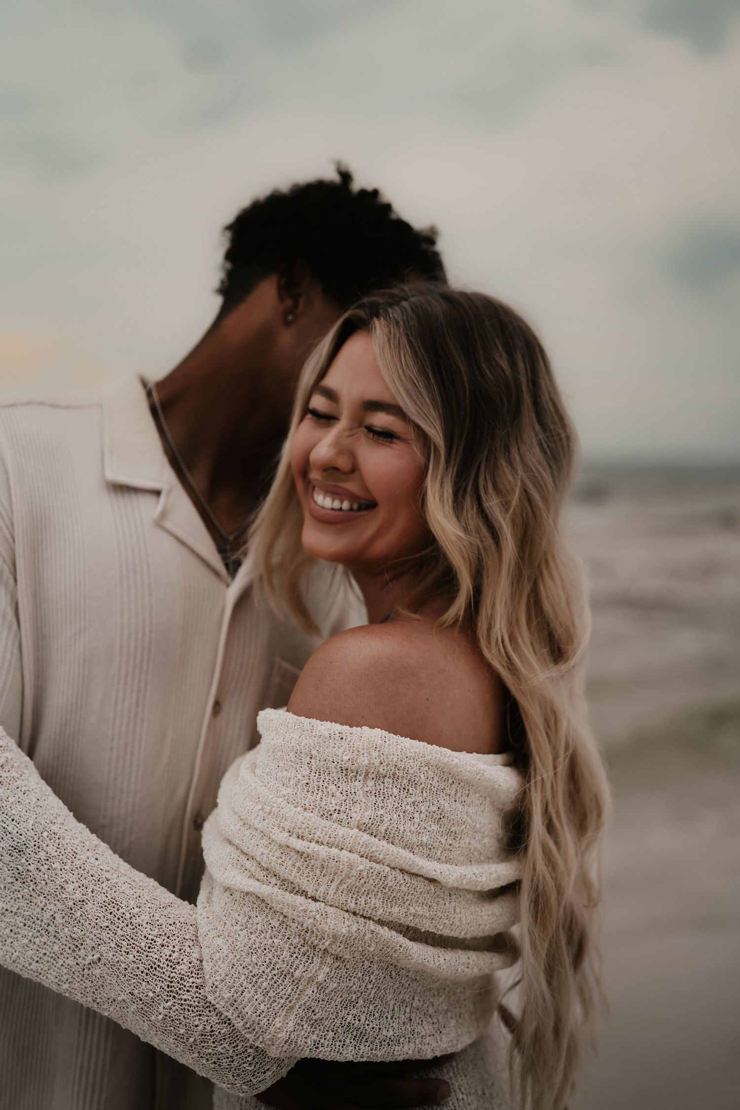 A smiling blonde woman in an off-shoulder sweater embraces her partner on a beach at dusk.